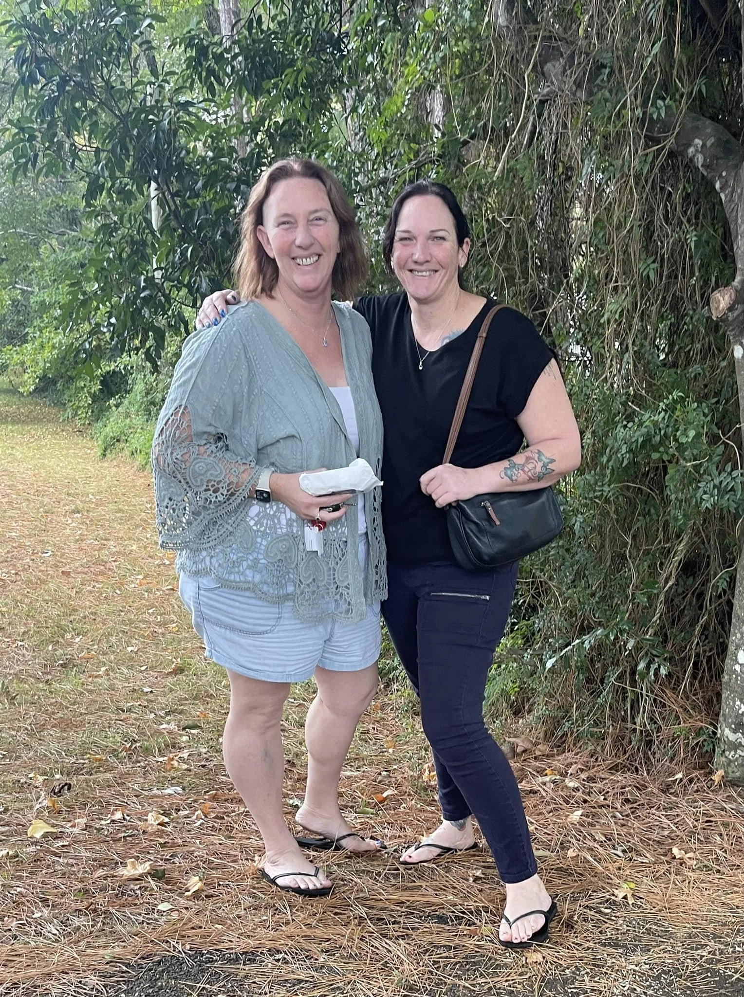 Two women standing outdoors on a dirt path, smiling and posing for the photo. One woman has shoulder-length brown hair and is wearing a light blue lace cover-up over a white top, white shorts, and flip-flops. The other woman has black hair tied back,