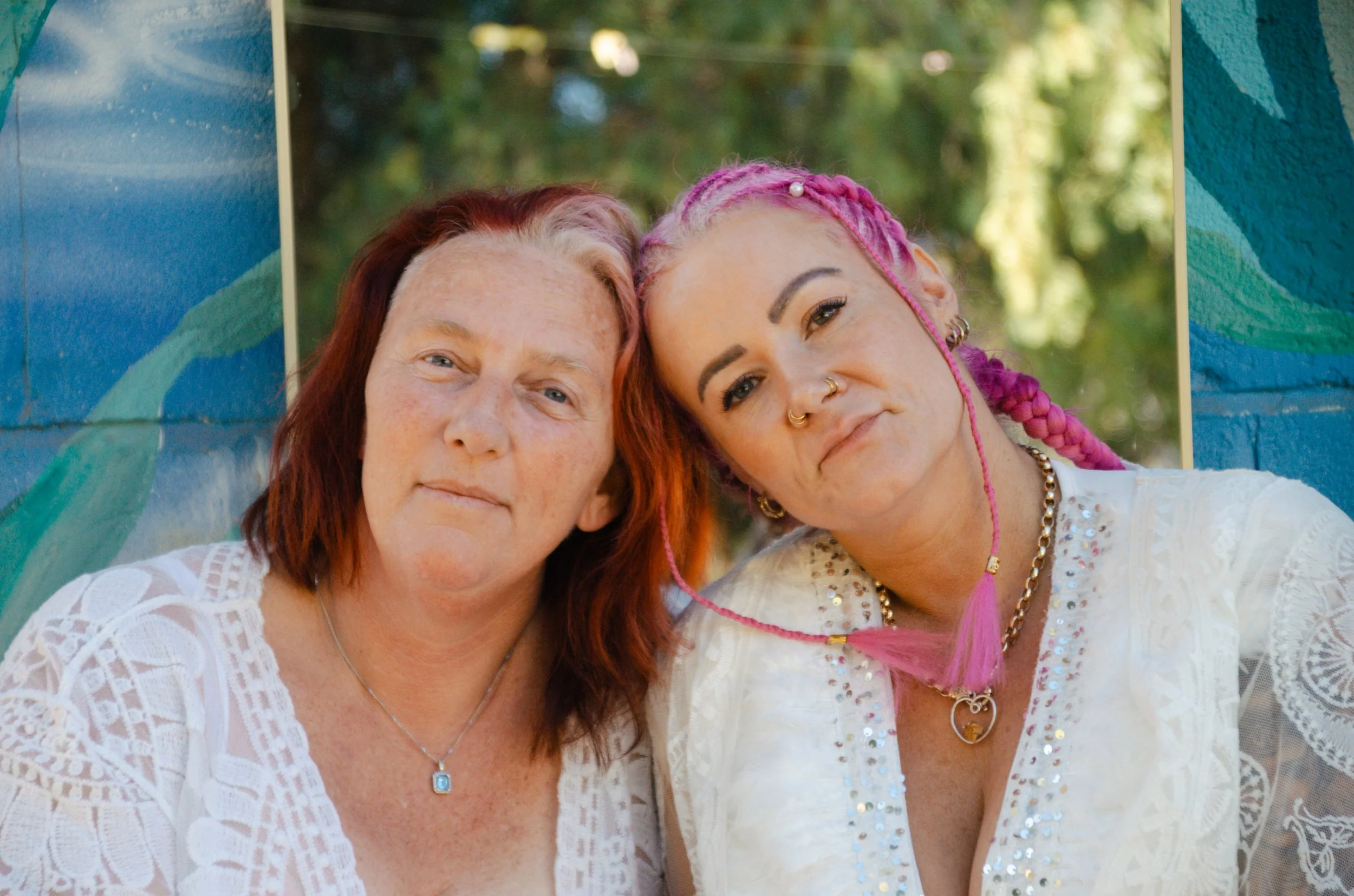 Two women with colorful hair and nose rings posing close together outdoors, with trees in the background.