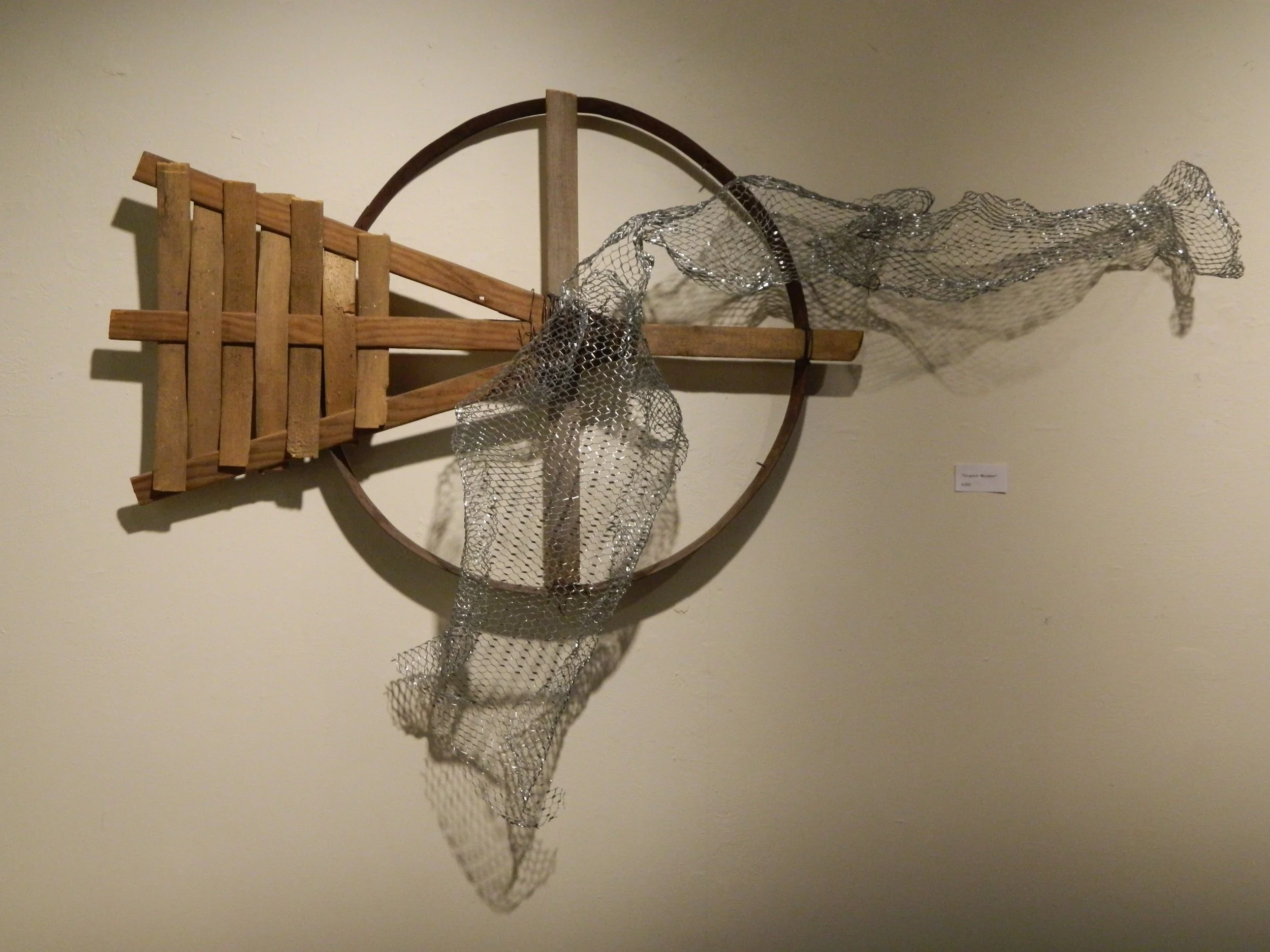 Mixed media art piece featuring a wooden wheel with a chainmail and wooden structure attached, displayed on a plain wall.