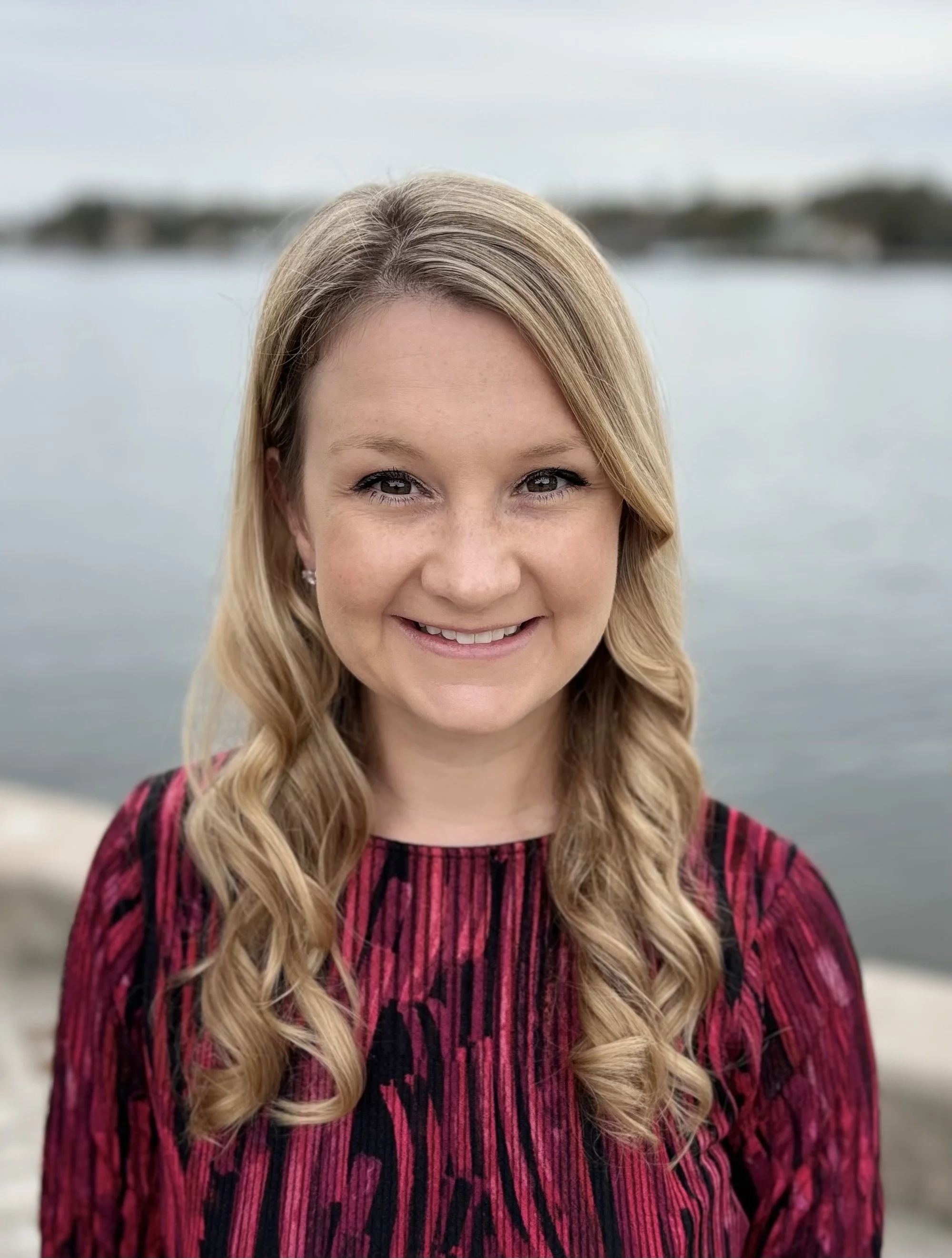 A woman with long blonde curly hair smiling outdoors near a body of water, wearing a red and black patterned top.