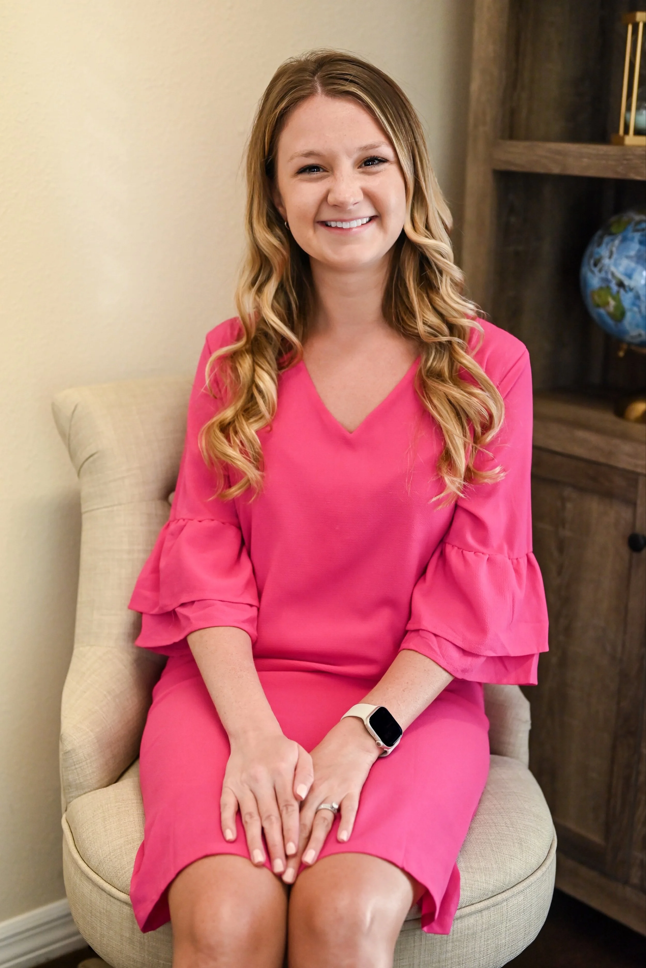 A young woman with long blonde curly hair sitting on a beige chair, smiling, wearing a pink dress with three-quarter length ruffled sleeves and a smartwatch on her left wrist, in a room with a wooden bookshelf and an Earth globe in the background.