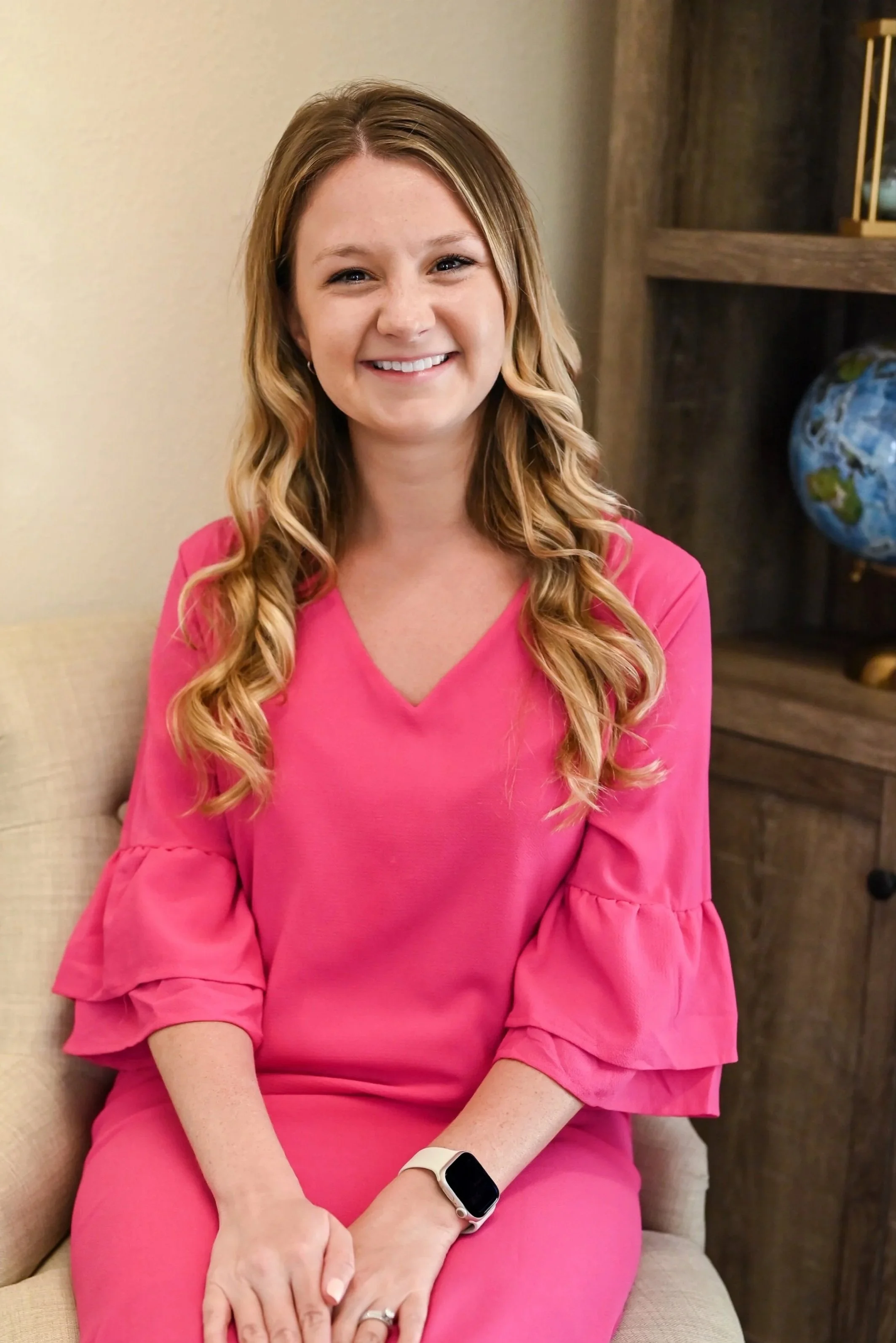 Young woman with long curly blonde hair wearing a pink dress with ruffled sleeves, sitting on a beige chair in front of a wooden shelf with a globe and decorative items, smiling at the camera.