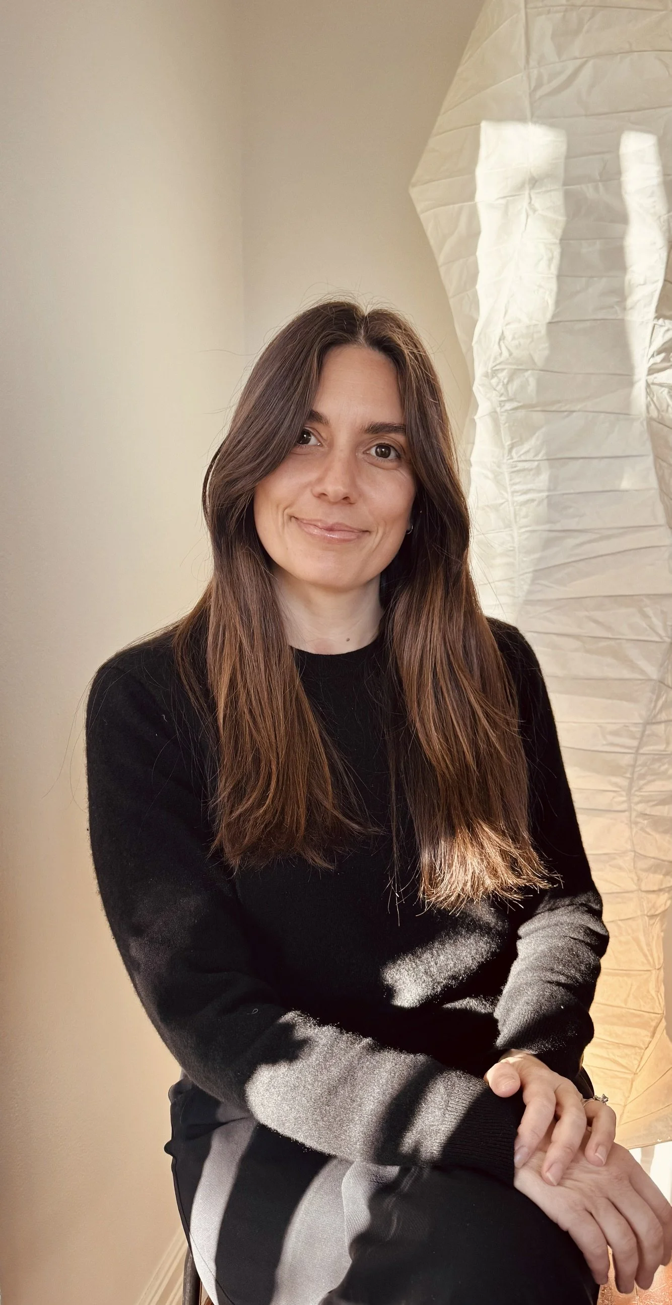 A woman with long brown hair, smiling, sitting with her hands clasped on her lap, wearing a black sweater and black and gray striped pants, in front of a beige wall and a white paper lantern.