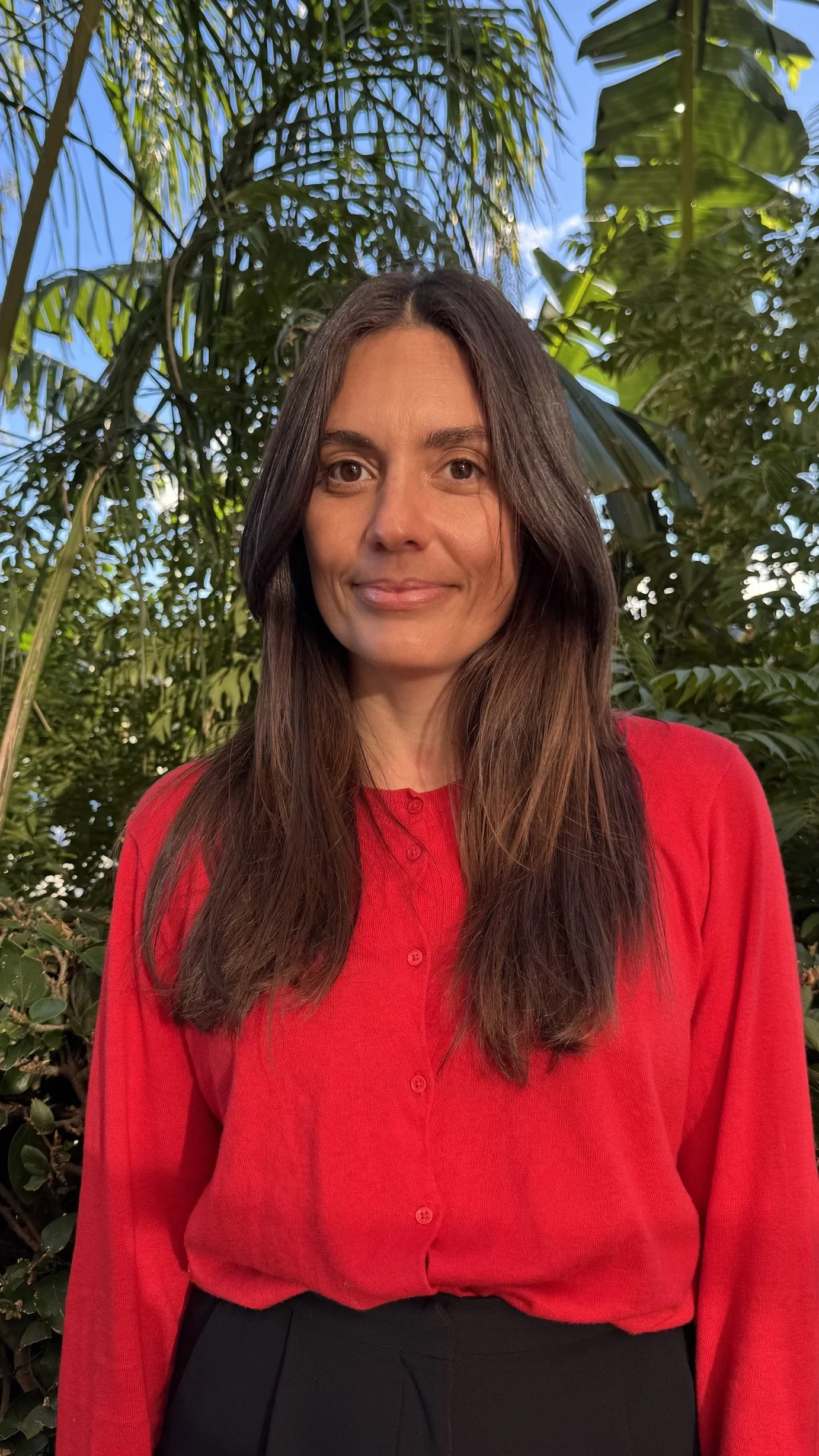 A woman with long dark hair and light skin wearing a red blouse and black pants, standing outdoors with lush green tropical plants and a clear blue sky in the background.