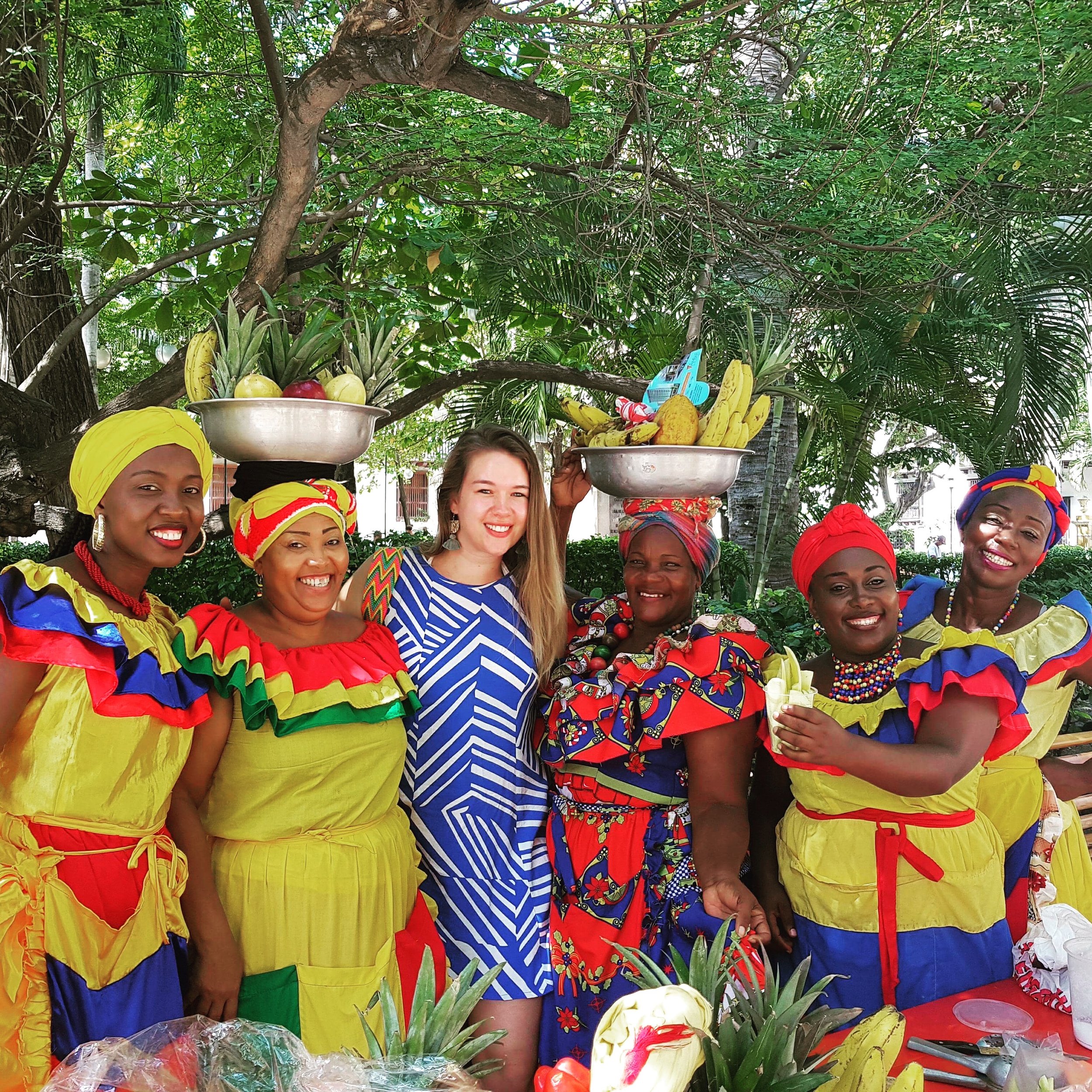 Group of women in colorful traditional outfits celebrating outdoors, with two women balancing baskets of fruit on their heads, surrounded by lush greenery.