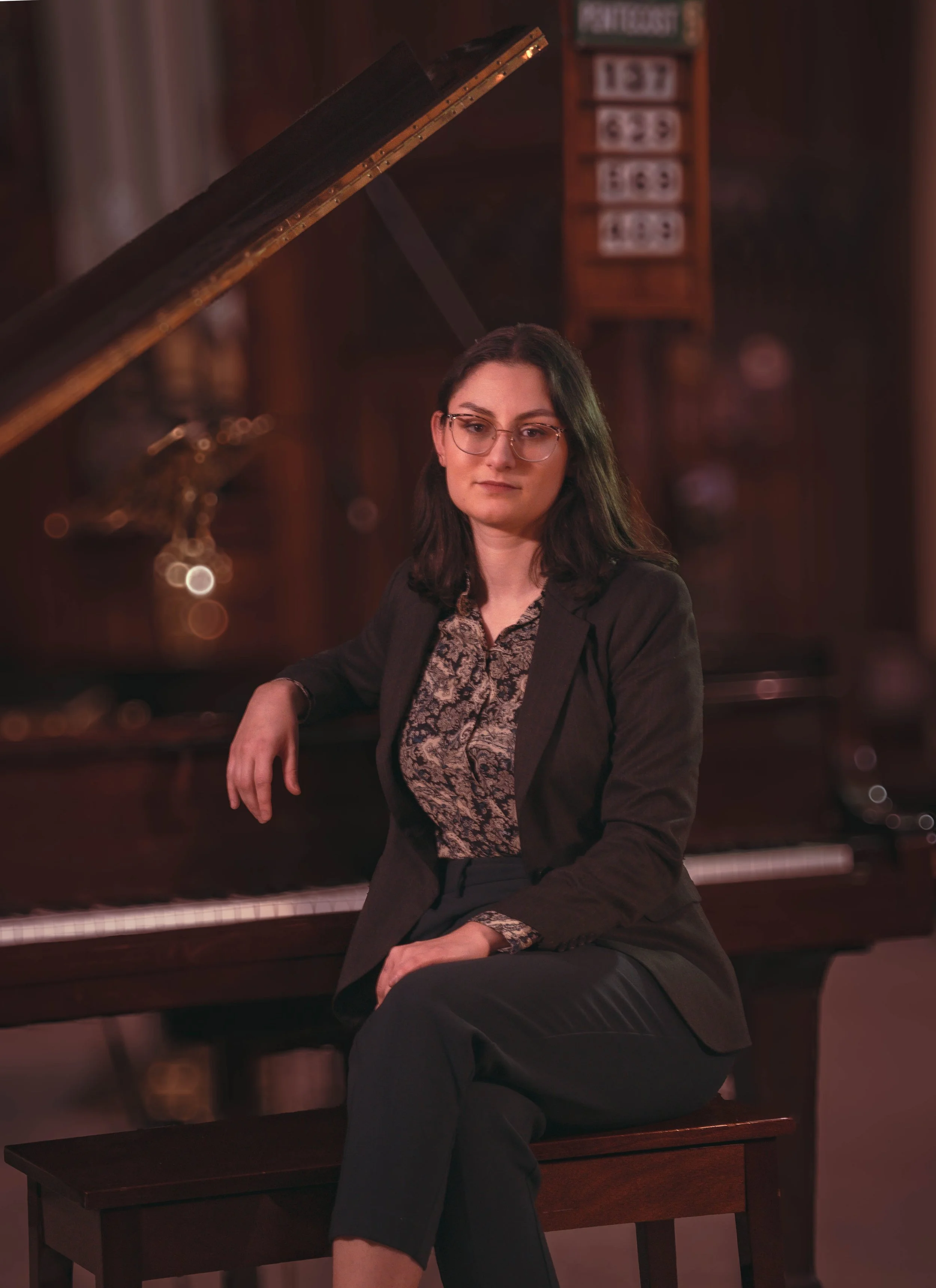 A woman with dark hair and glasses sitting on a bench beside a grand piano in a dimly lit room with wood walls, a bulletin board, and musical equipment.
