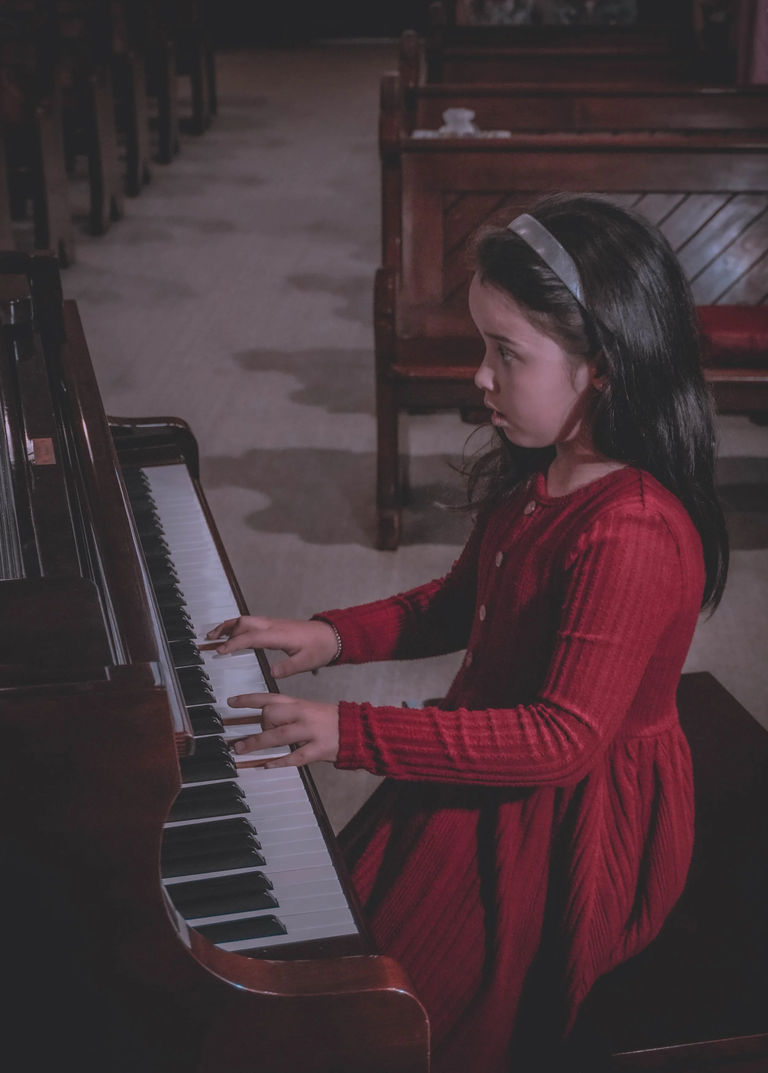 Young girl in a red dress playing the piano in a dimly lit room with wooden pews in the background.