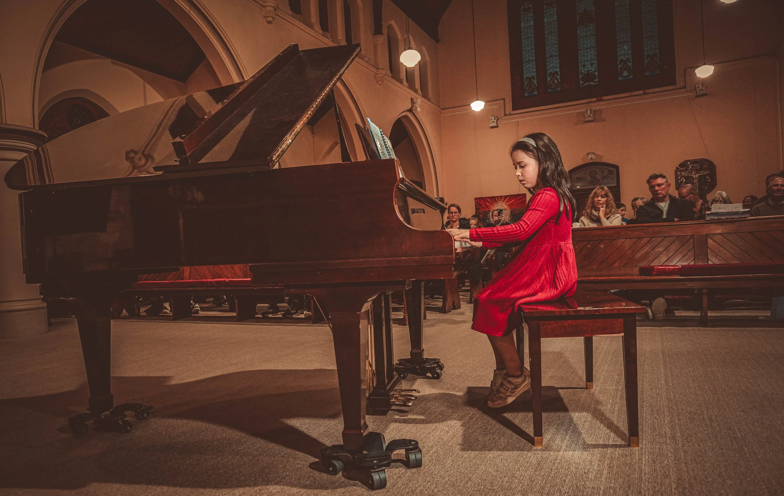A young girl in a red dress playing piano in a church or concert hall, with an audience watching her.