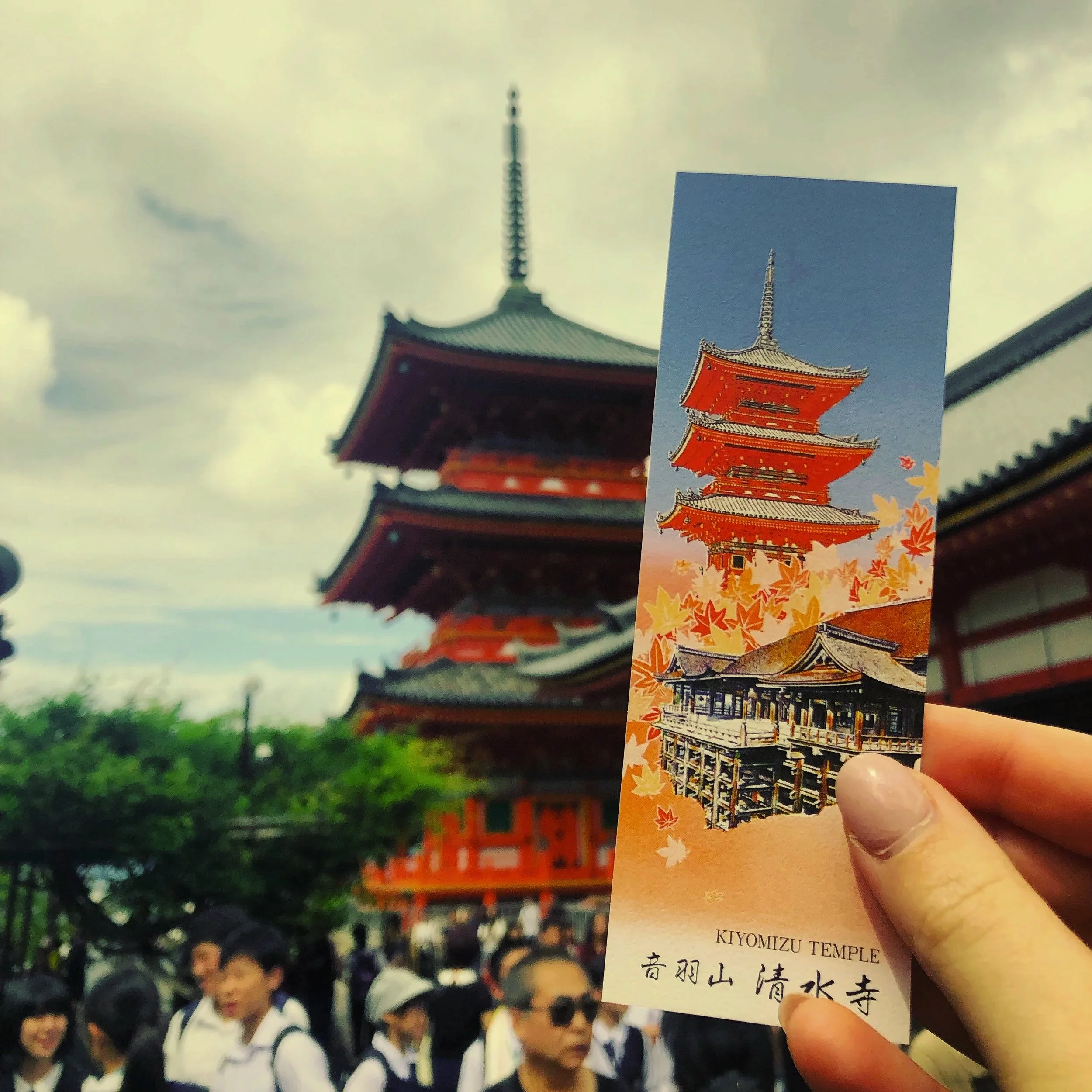Tourist holding a souvenir ticket with an illustration of Kiyomizu Temple, with the actual multi-tiered pagoda in the background surrounded by people and trees.