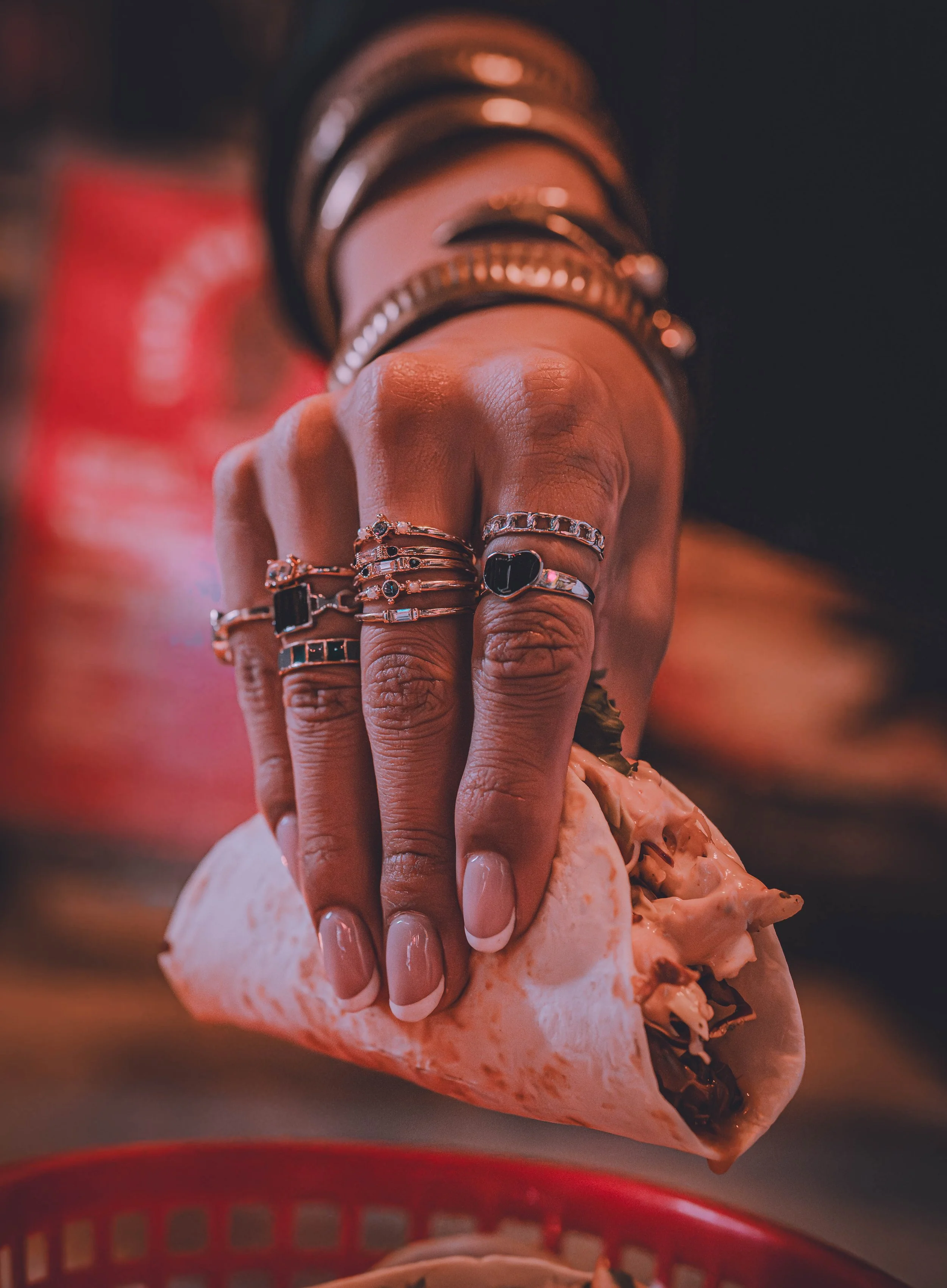 A person's hand with multiple silver rings holding a taco with meat and vegetables, over a red basket, in a low-light setting.