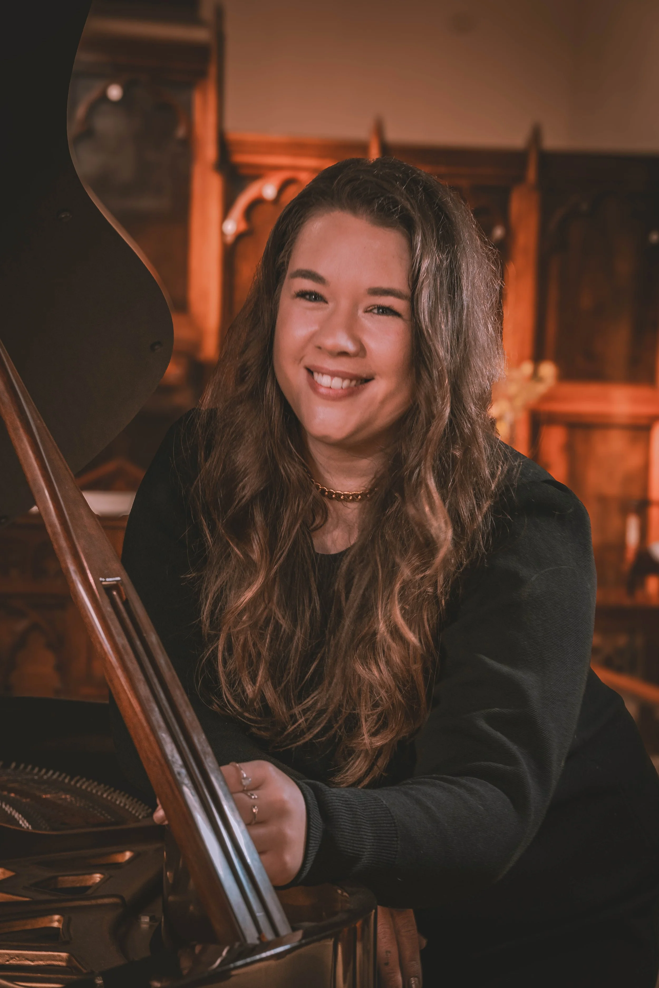A young woman with long wavy brown hair, smiling, sitting at a grand piano with the lid open, inside a wood-paneled room with ornate woodwork.