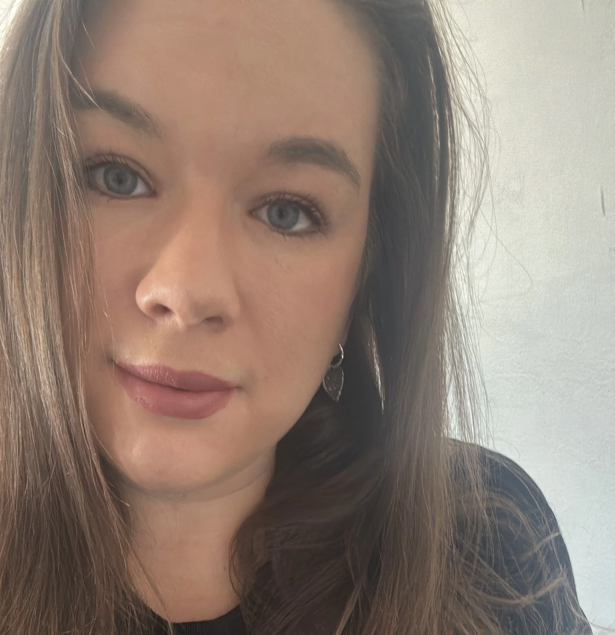 Close-up of a young woman with long brown hair, blue eyes, wearing silver earrings and a black top, against a neutral background.