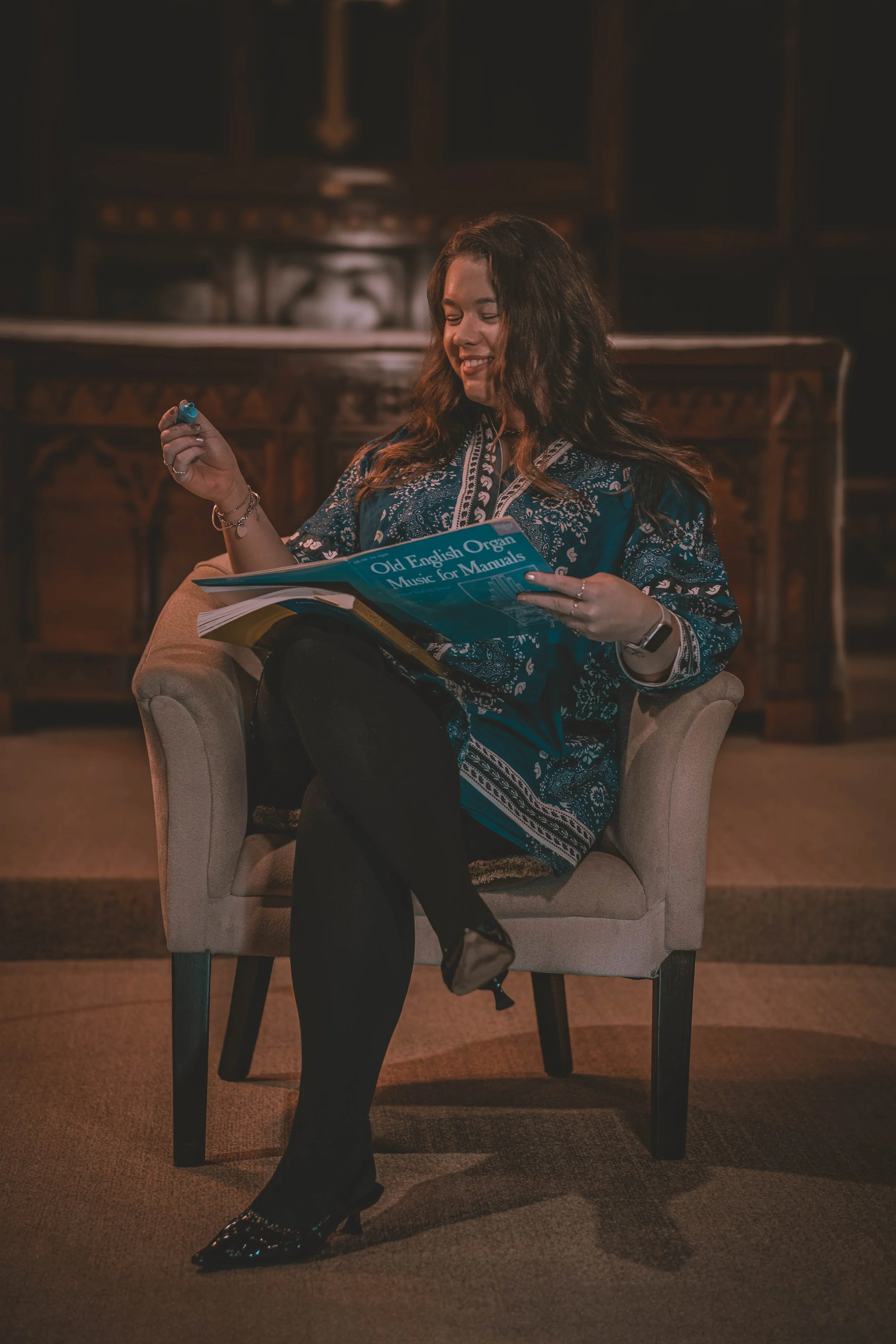 Woman sitting on an armchair, reading music for manual organ plays in a room with wooden furniture. She is smiling and holding a pen while looking at a music book titled 'Old English Organ Music for Manuals'.