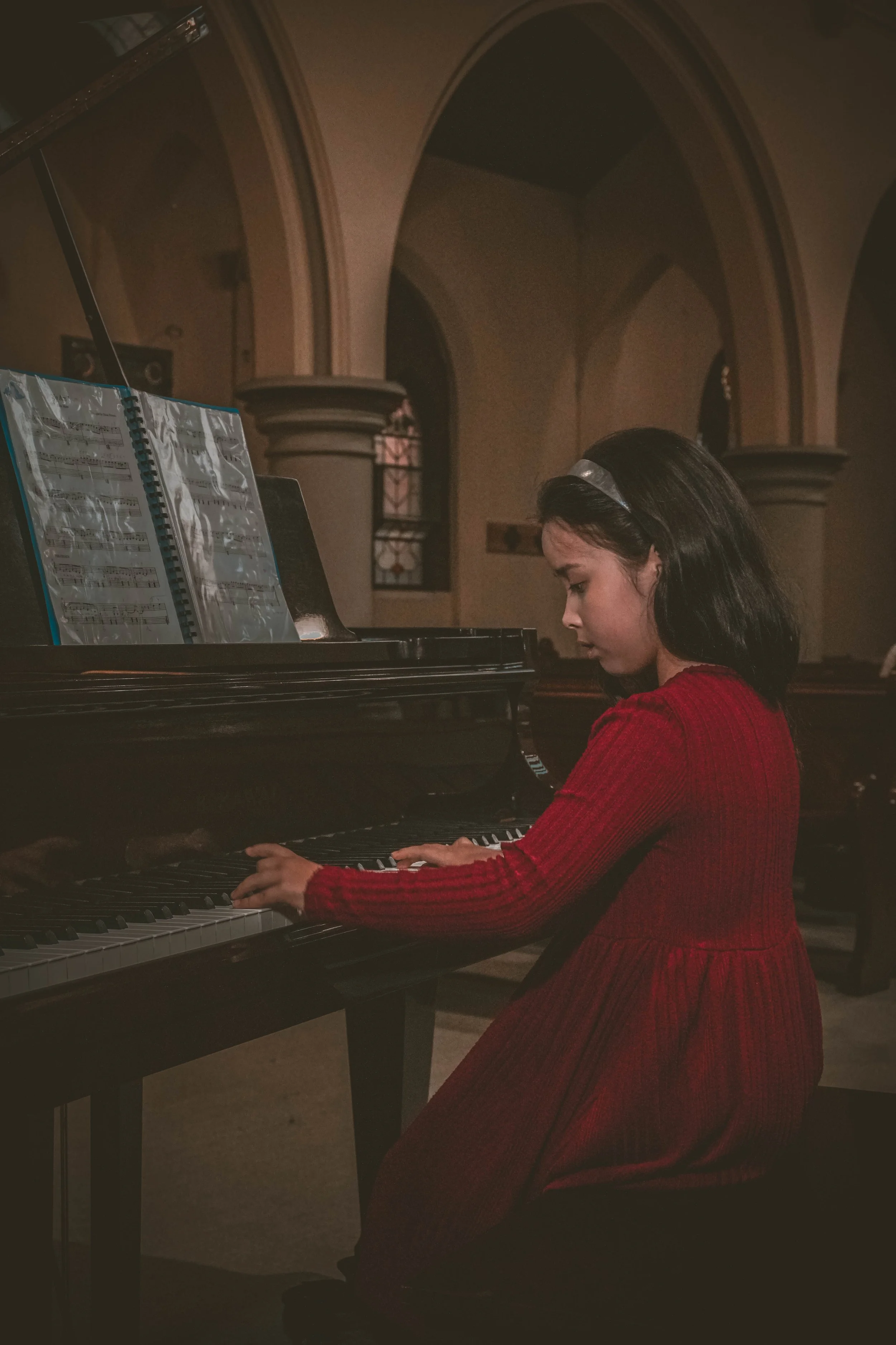 A young girl in a red dress playing the piano in a dimly lit room with arched windows and classical architecture.
