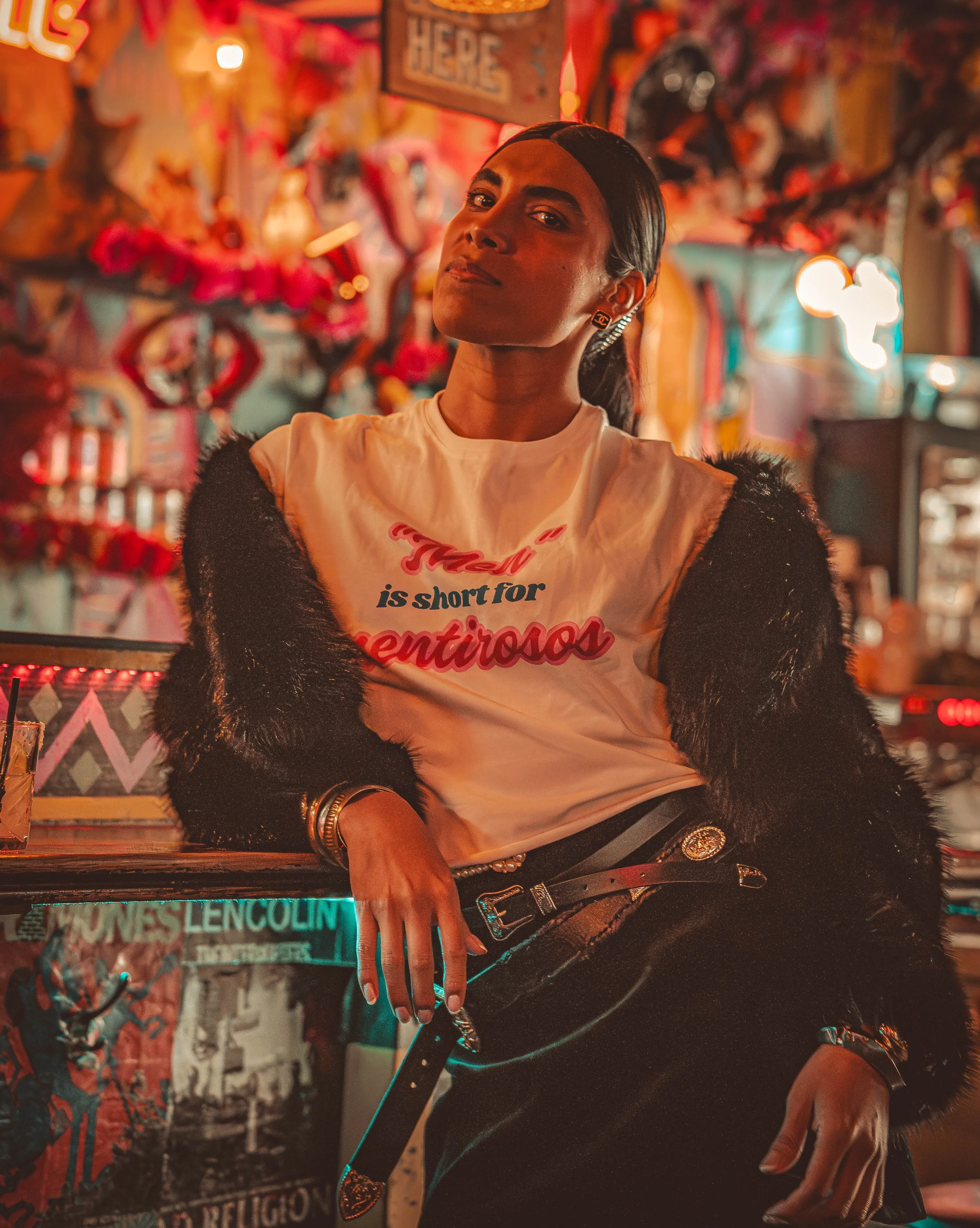 A woman with dark hair styled in a sleek ponytail, wearing earrings, a white graphic T-shirt, black pants with a belt, and a black furry jacket, sitting indoors against a vibrant, colorful background with illuminated signs and wall art.