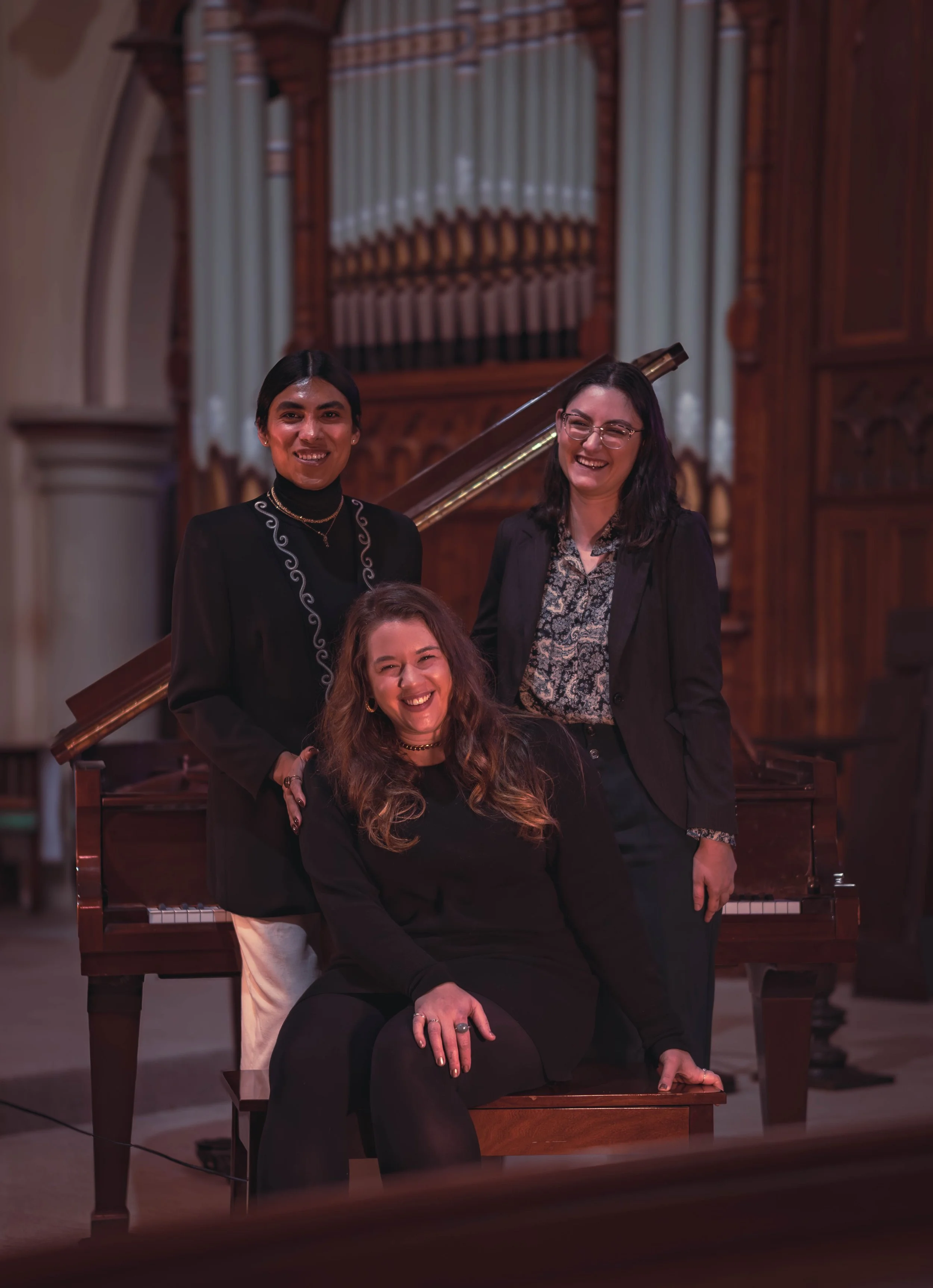Three women smiling inside a church with a wooden pipe organ in the background, one sitting on a bench and two standing behind her.