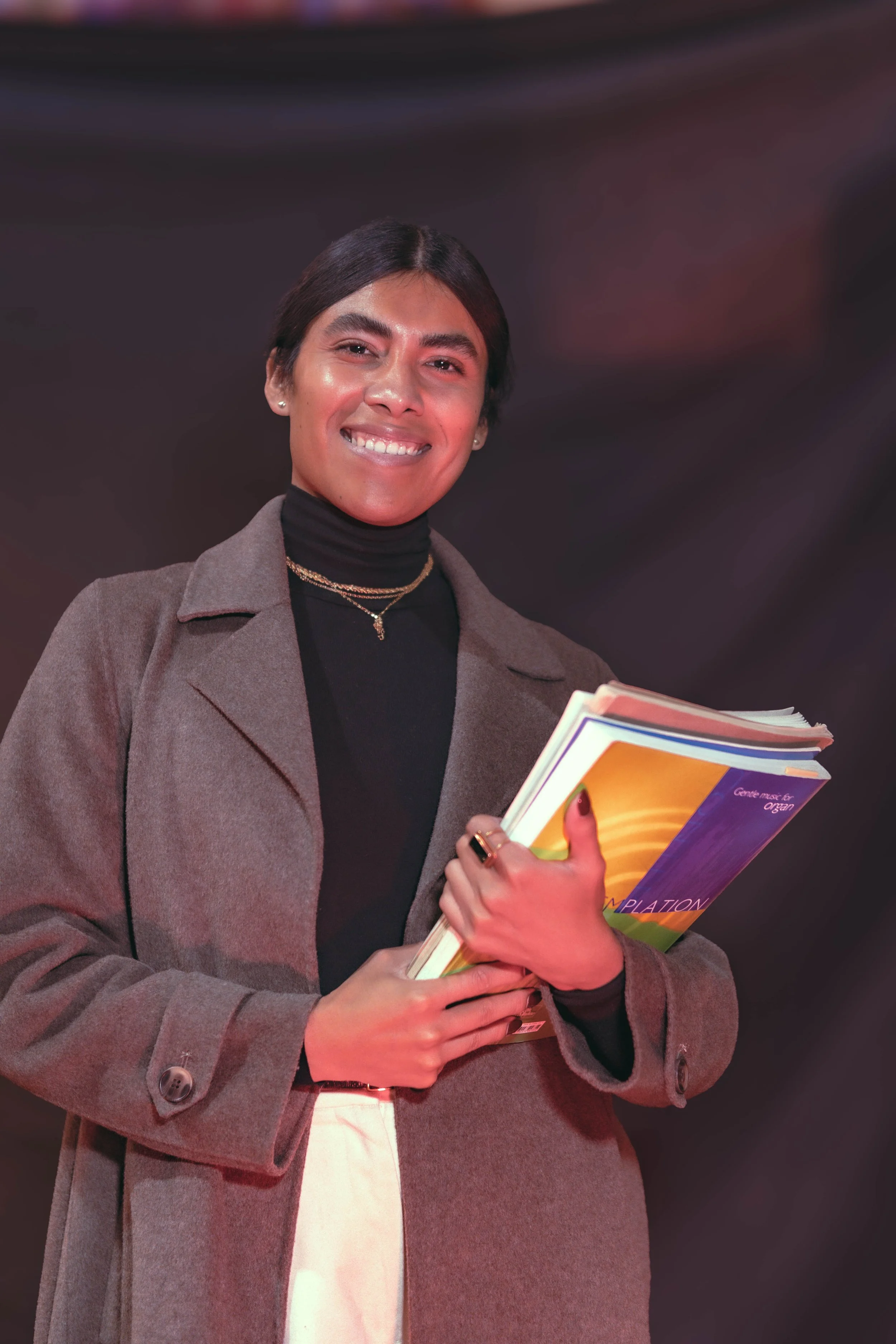 A woman with short dark hair smiling at the camera, wearing a gray coat over a black turtleneck, holding several colorful books or notebooks in her hand against a dark blurred background.