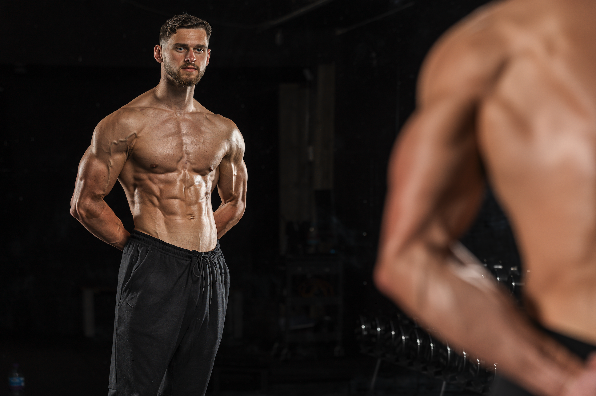 A muscular young man with short brown hair and a beard, standing shirtless and wearing black athletic shorts, appears to be in a gym. He has a serious expression and his hands are clasped behind his back. In the foreground, there is a blurred part of another person, also shirtless, possibly a woman, with a muscular physique.