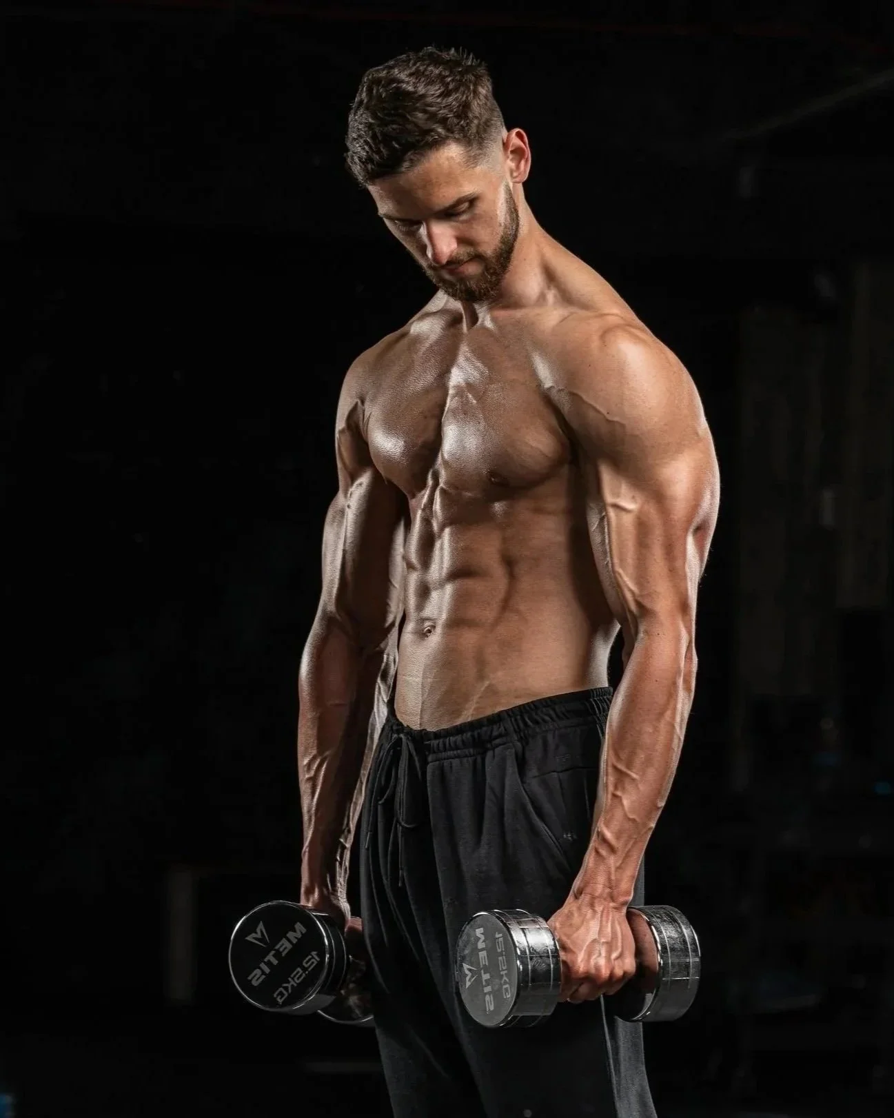 A shirtless man with a muscular physique lifting dumbbells in a dark gym setting.