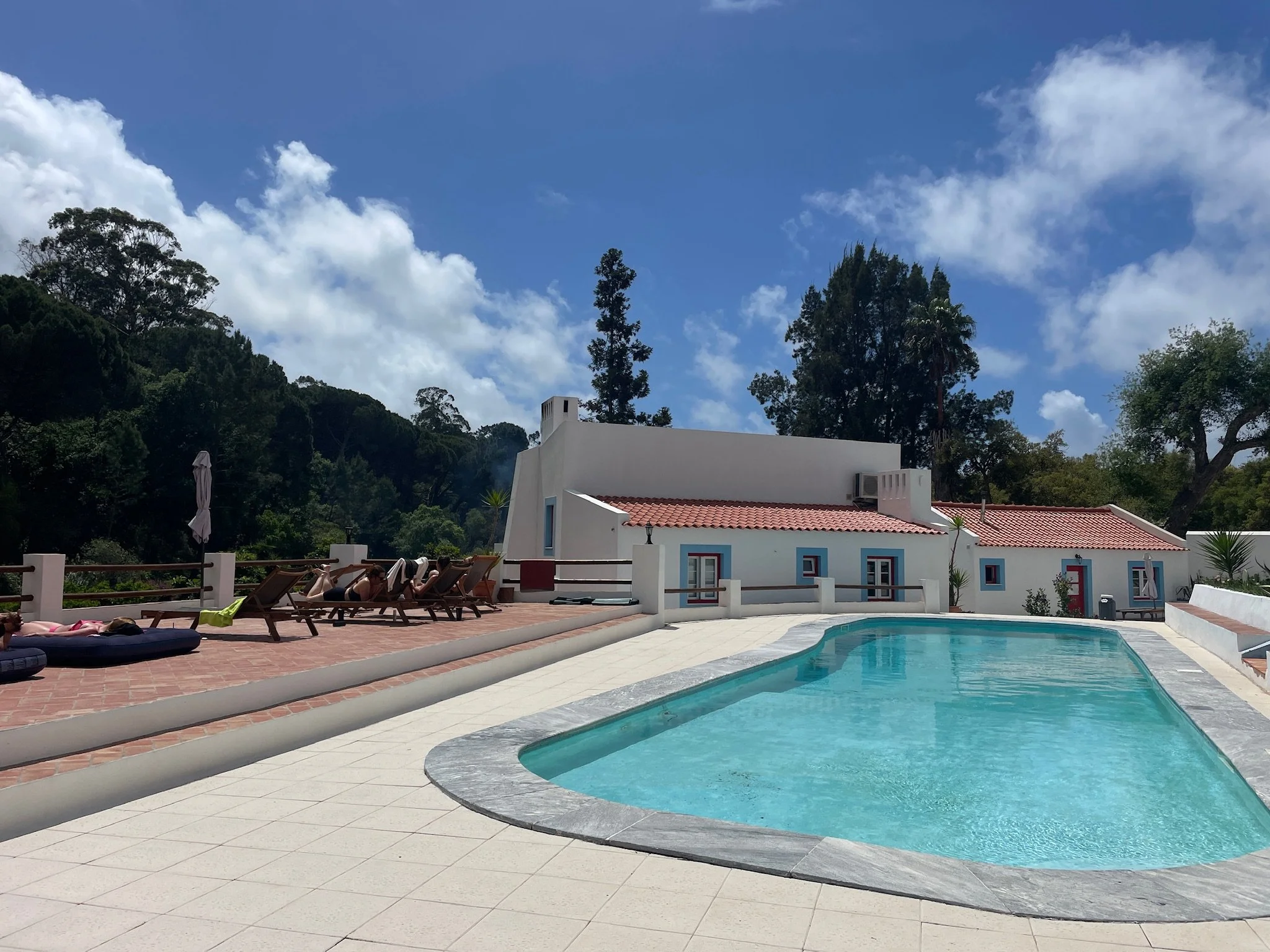 A swimming pool with poolside lounge chairs and umbrellas in front of a white building with a red-tiled roof, surrounded by trees and a bright blue sky with clouds.
