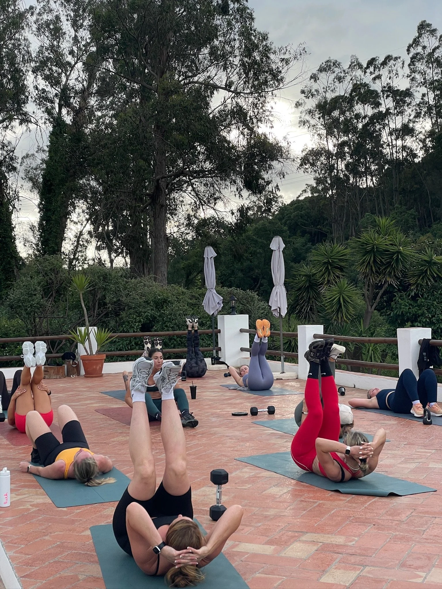Group of women participating in an outdoor fitness class, lying on exercise mats and lifting their legs with hands behind heads, with trees in the background.