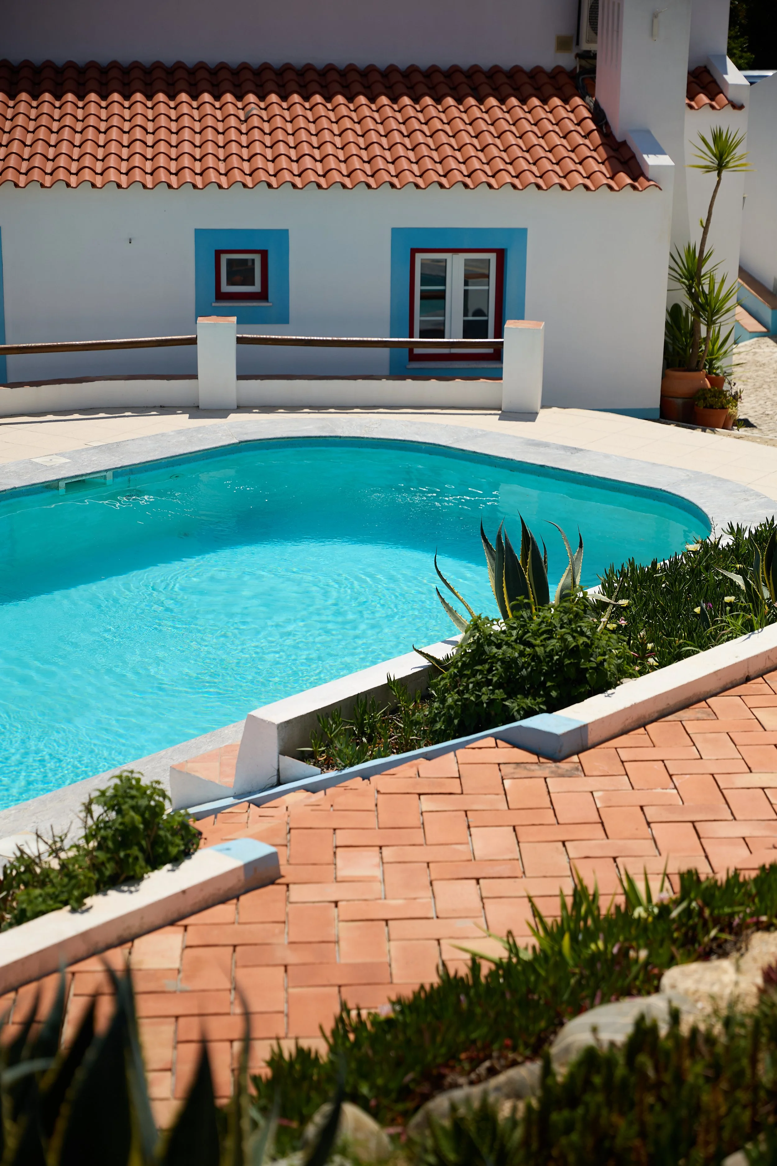 A swimming pool with turquoise water in front of a white house with a red-tiled roof, surrounded by plants and a brick patio.