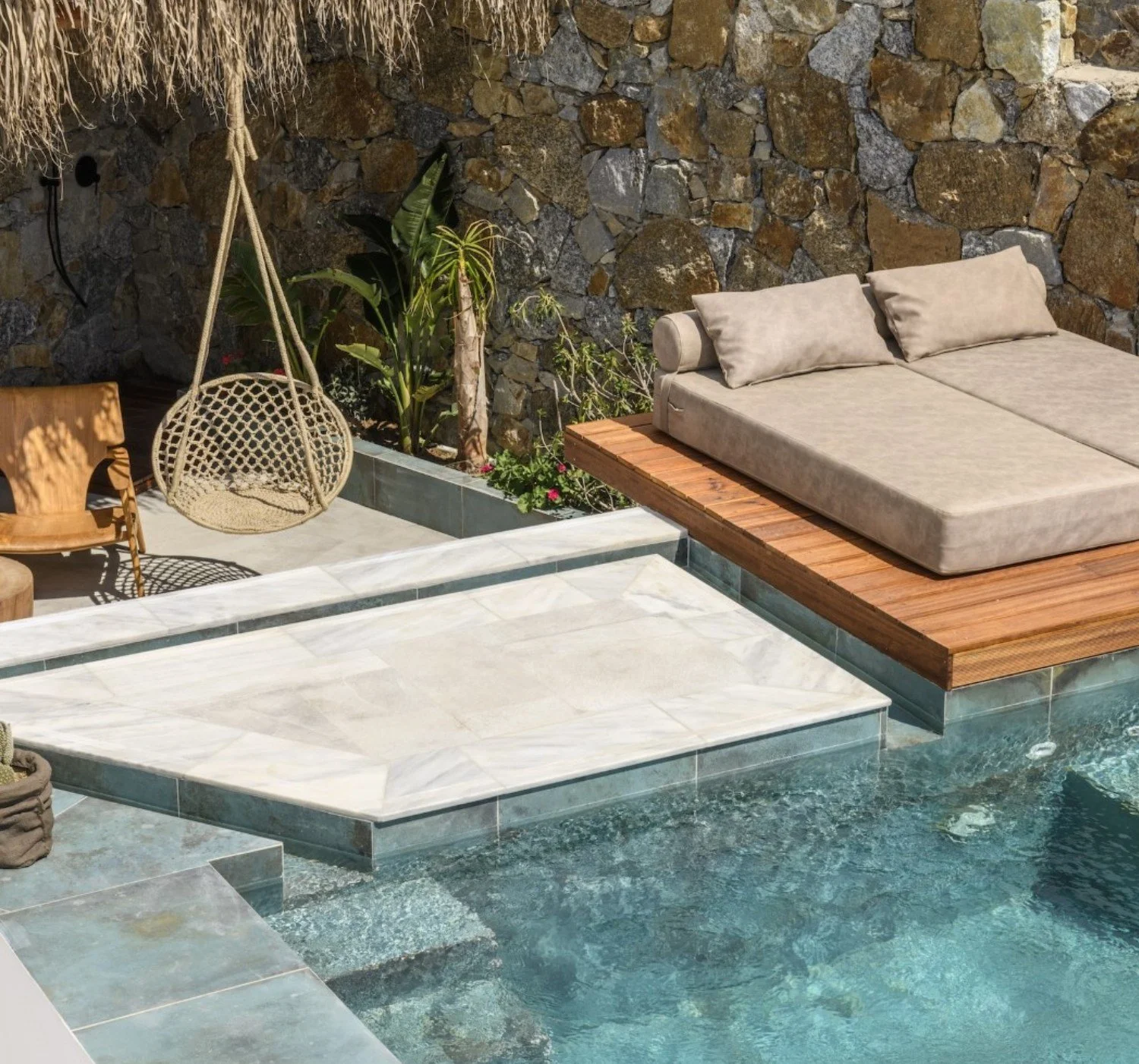 Outdoor pool area with a stone wall background, two lounge chairs with cushions on a wooden platform, a hanging wicker chair, and plants.