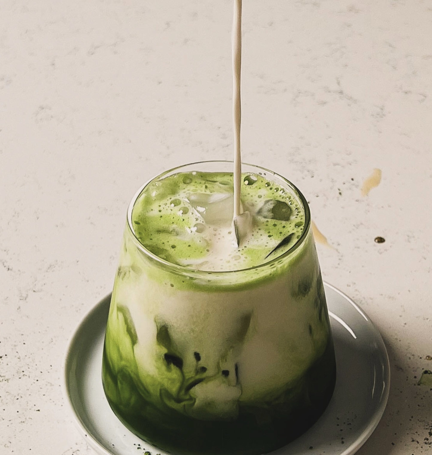 A glass with a green and white swirl pattern, filled with a beverage and ice cubes, placed on a small white plate, with milk being poured into it.