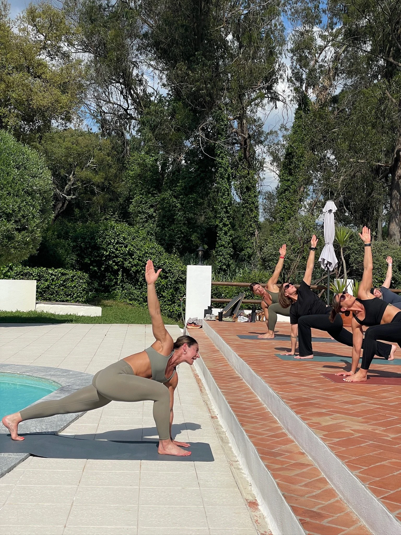 Group of women practicing yoga outdoors by a swimming pool, in a Warrior pose, with trees and cloudy sky in the background.