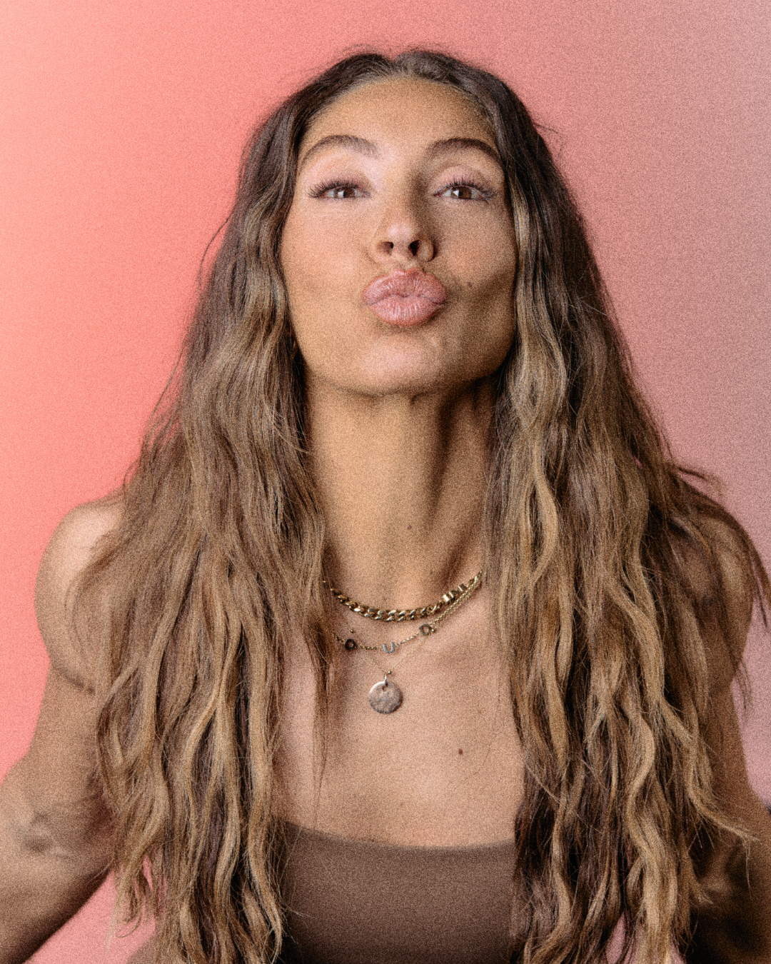 A smiling woman in athletic wear posing in a gym with dark walls, a neon sign, and workout equipment in the background.