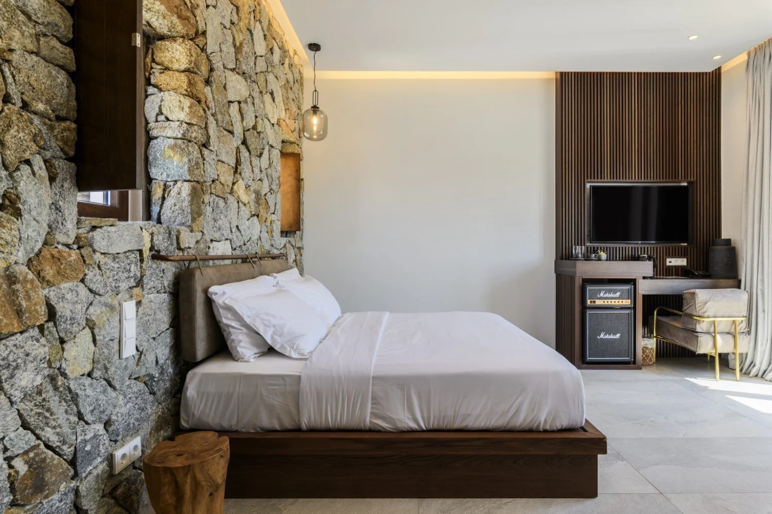 Modern hotel room with a stone accent wall behind the bed, a wooden bed frame, white bedding, a hanging pendant light, a flat-screen TV mounted on a wooden slat wall, a Marshall amplifier, a chair with gold legs, and floor-to-ceiling curtains.