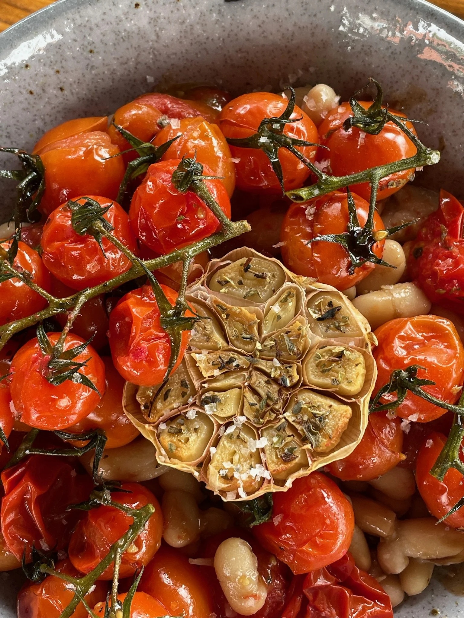 Cherry tomatoes on the vine, roasted with garlic, basil, sea salt, and herbs, with a halved garlic bulb in the center.