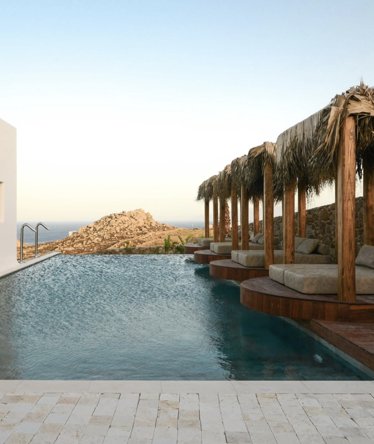 Infinity pool with poolside cabanas featuring straw thatched roofs overlooking a rocky hillside and ocean in the distance.