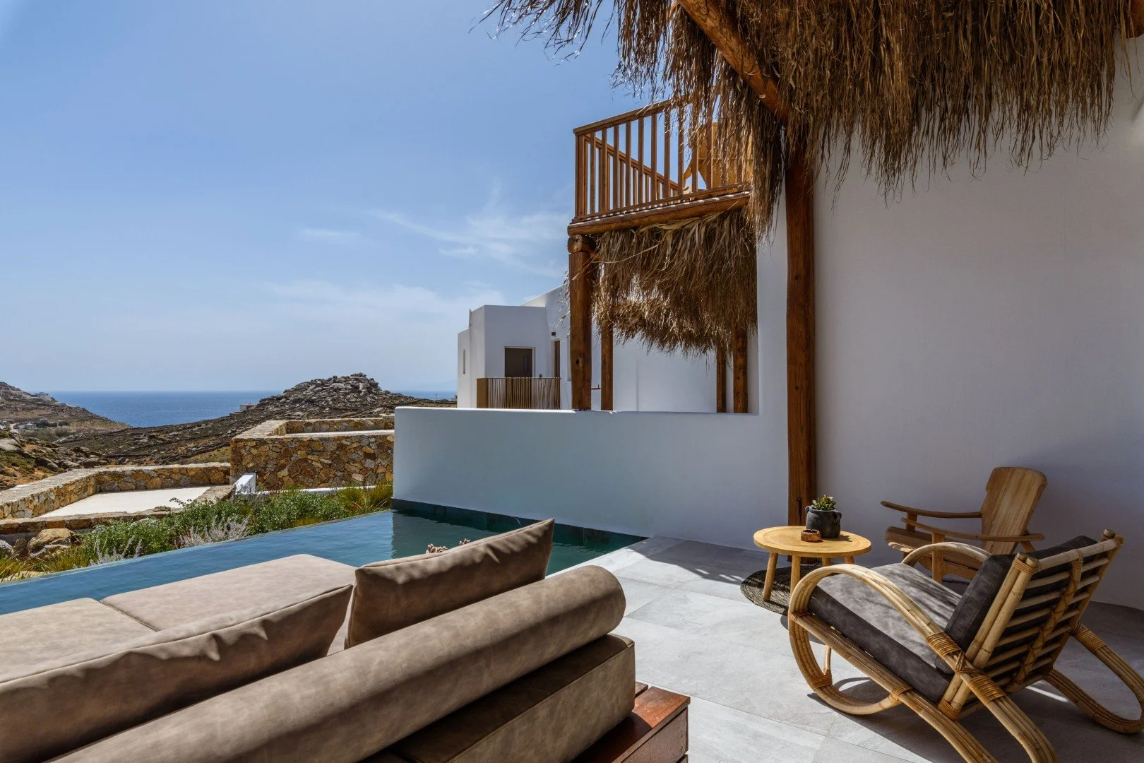 Outdoor patio with lounge chairs, small table, and pool overlooking rocky landscape and ocean in the distance, with thatched roof structure and white building