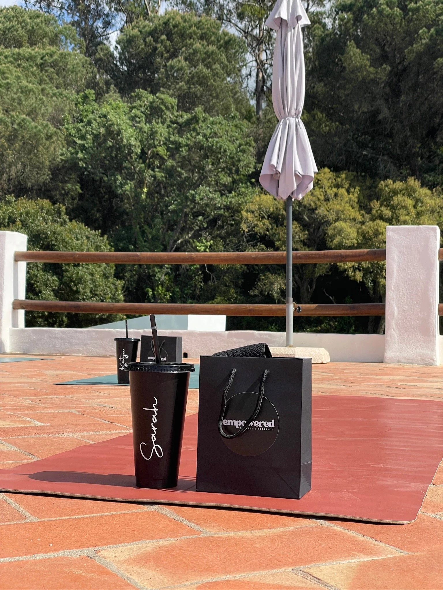 A black cup with the name 'Sparsh' written on it, a black gift bag with the 'Empowered' logo, and a black straw on a red mat on a brick patio. An outdoor area with trees and a white wall with a wooden rail and an umbrella in the background.