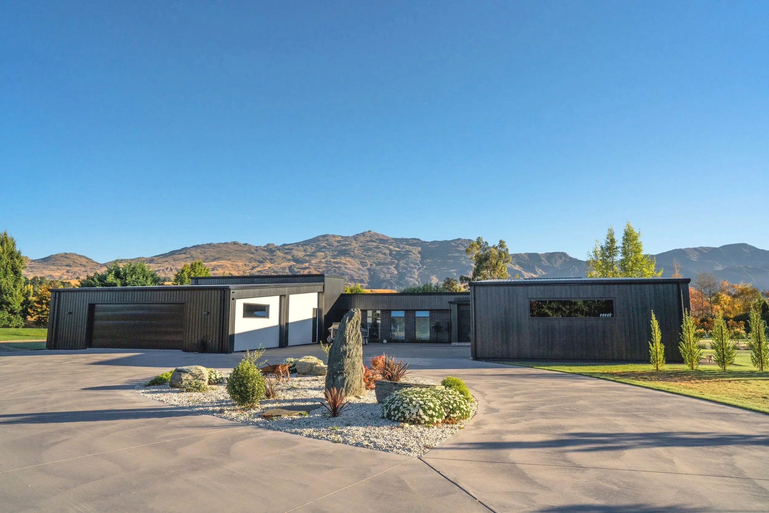 Modern black metal house with landscaped front yard, rocks, and plants, set against a mountain background under a clear blue sky.