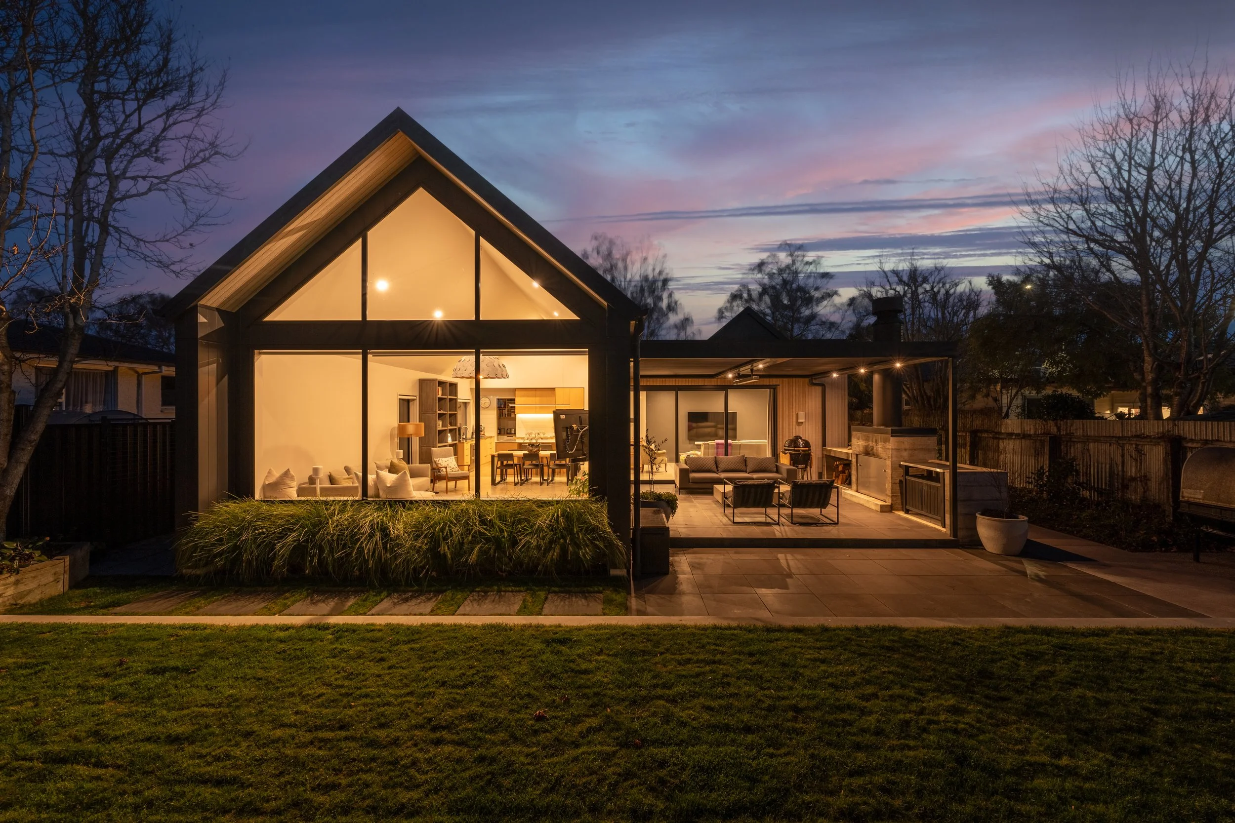 A cozy, illuminated house with large glass windows at dusk, showing a living room and outdoor patio with seating and a barbecue grill.