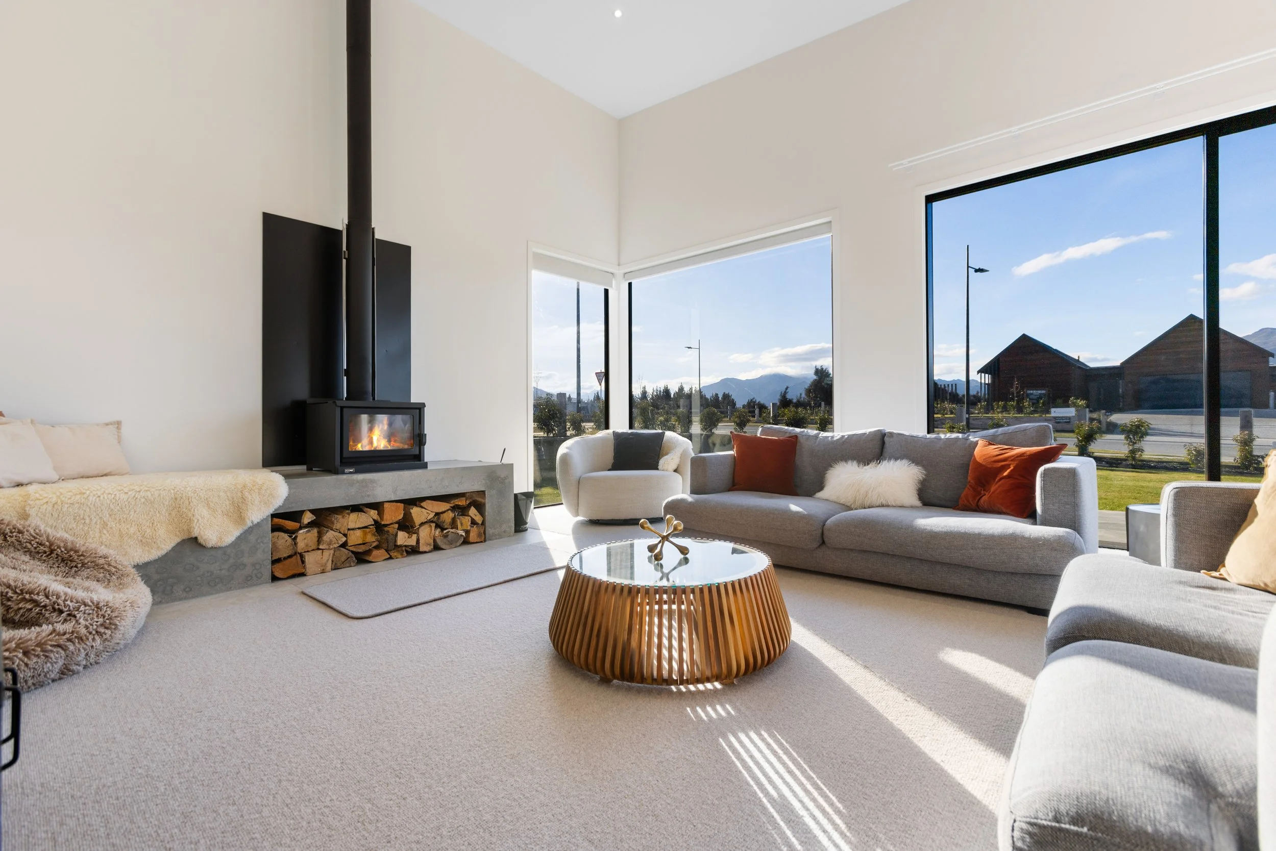 Sunlit living room with large windows, contemporary gray sofas with pillows, a glass-topped wooden coffee table, a white armchair, a modern wood stove with fire, and a view of mountains and houses outside.