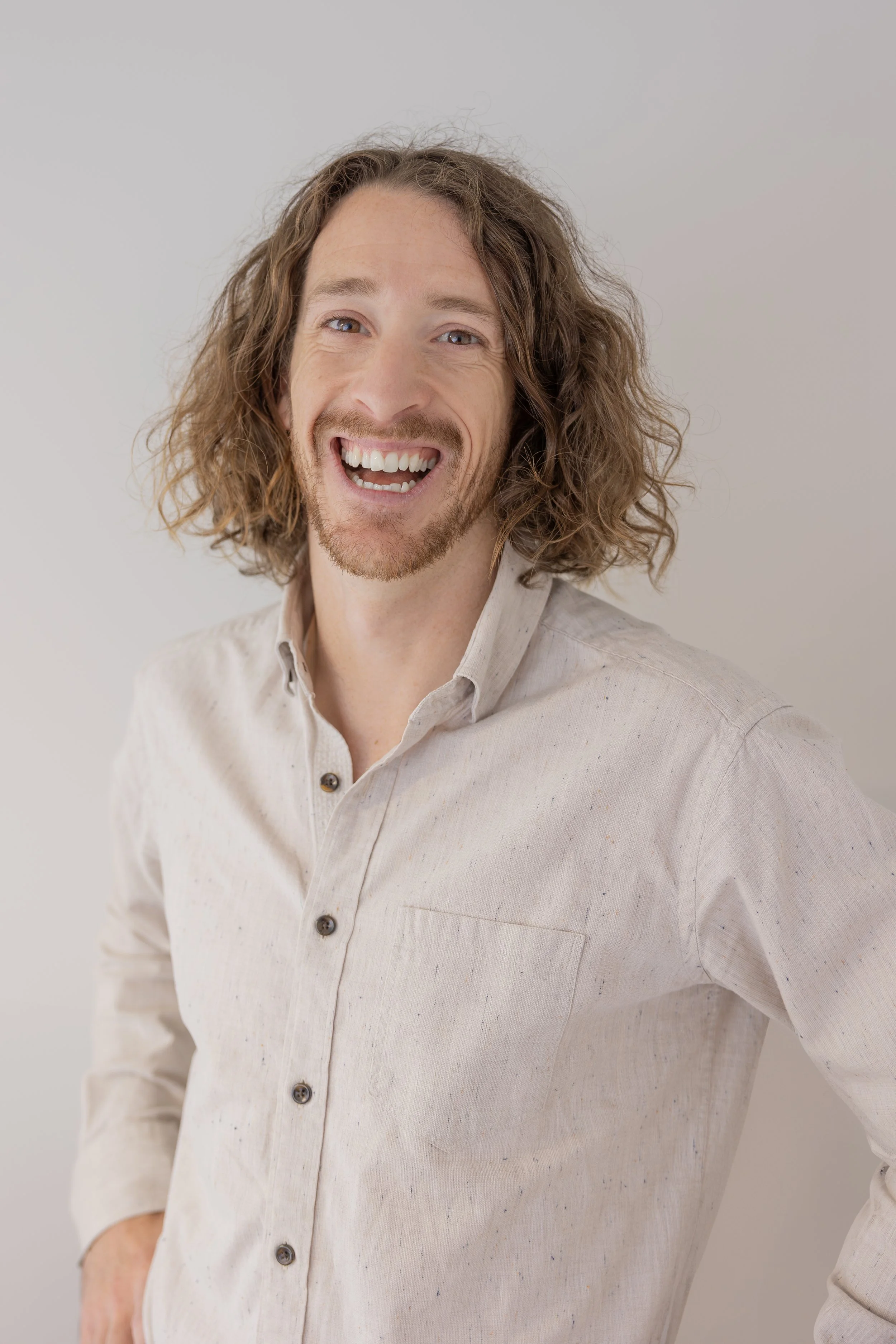 Smiling man with shoulder-length curly hair and a beard, wearing a light-colored button-up shirt, standing against a plain light background.