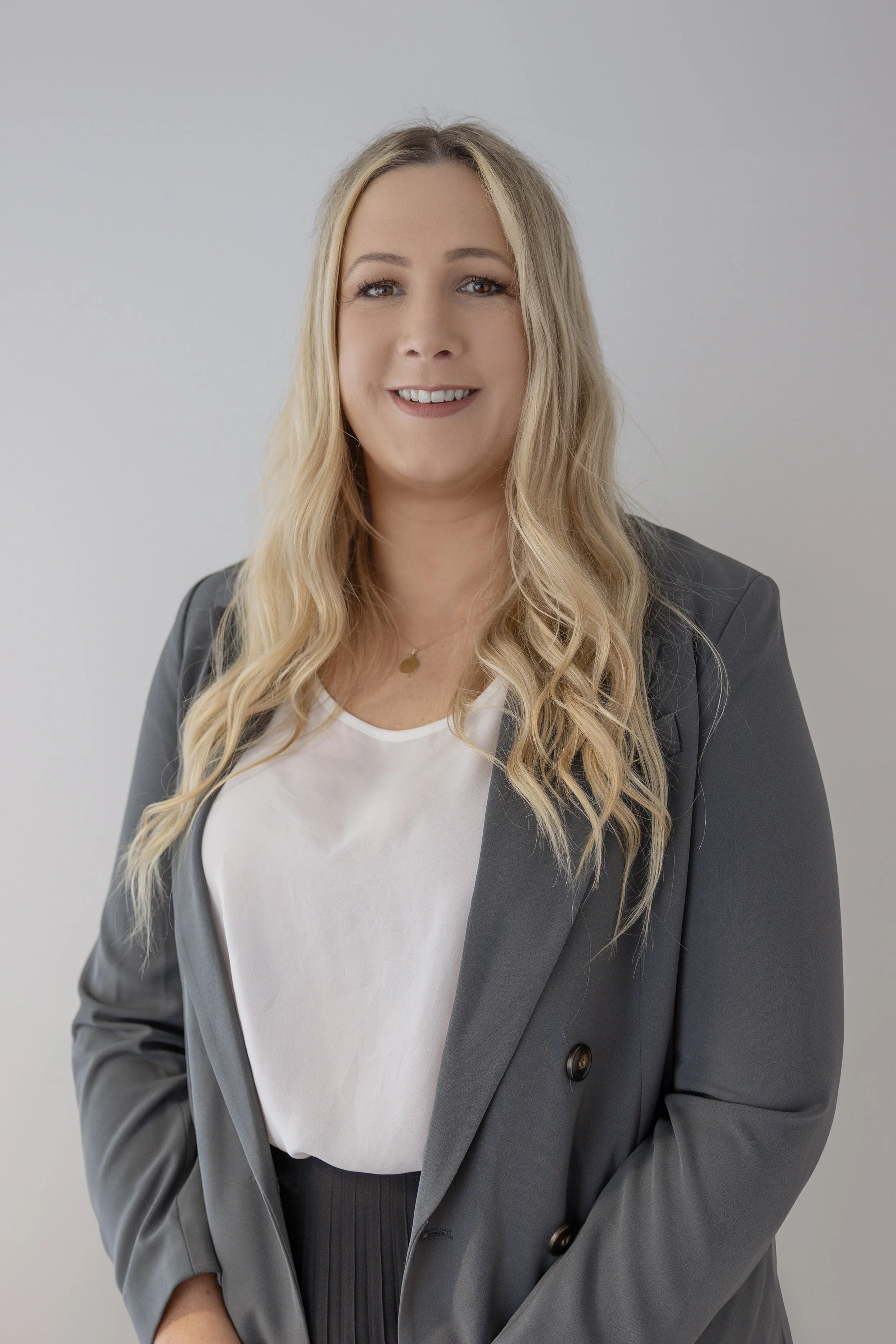 A woman with long blonde hair smiles at the camera while wearing a gray blazer over a white blouse, standing against a plain light gray background.