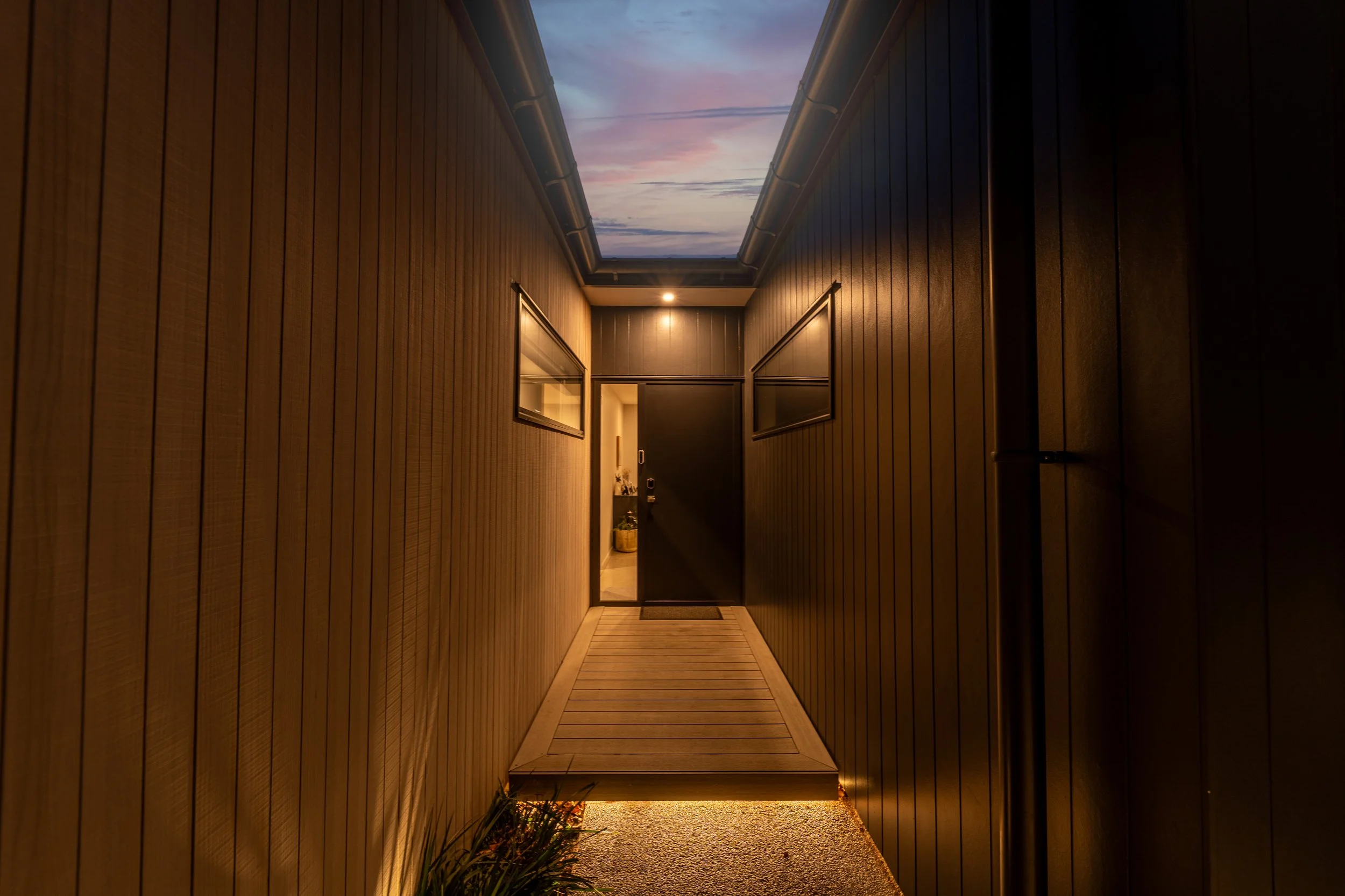 A narrow outdoor corridor with wooden panel walls and a wooden ramp leading to a front door, illuminated by warm lights with a sky view through a skylight ceiling.