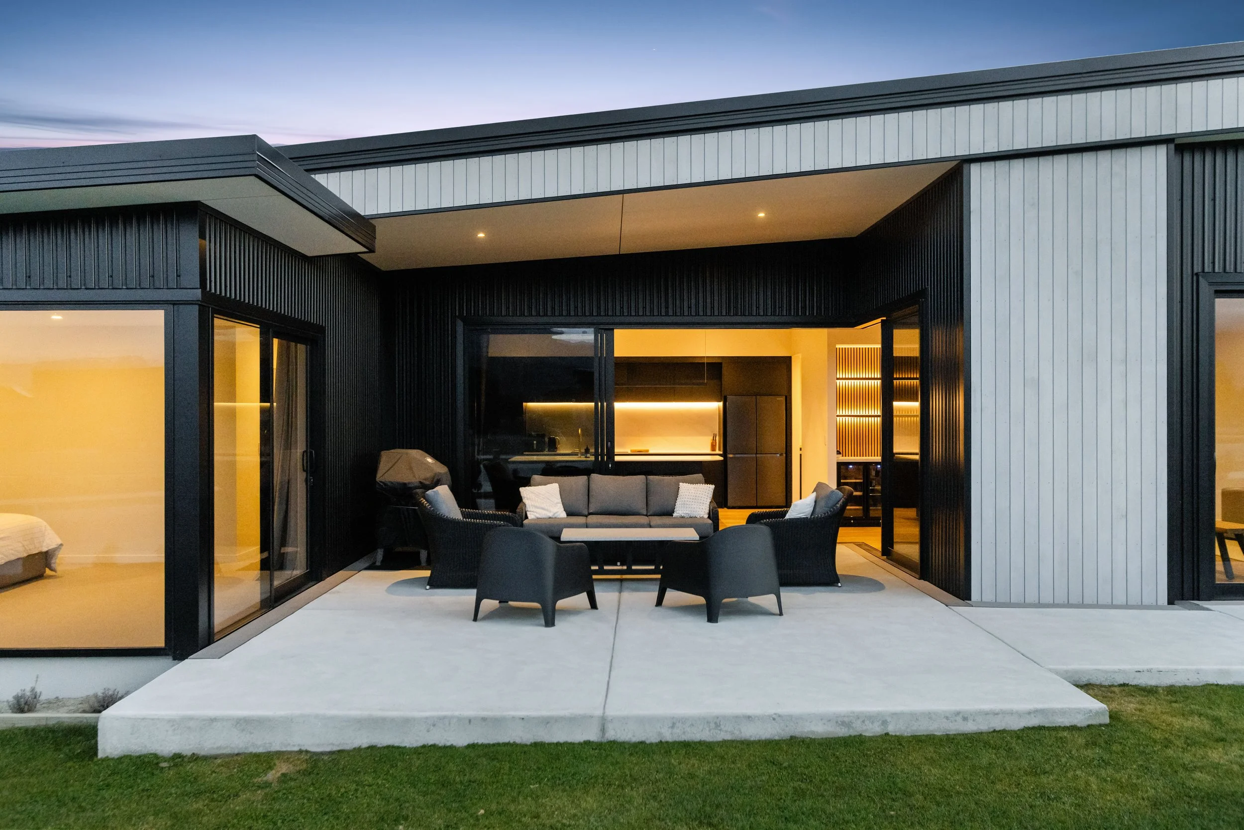 Modern house with large patio, outdoor seating, and visible interior kitchen and bedroom, during dusk.