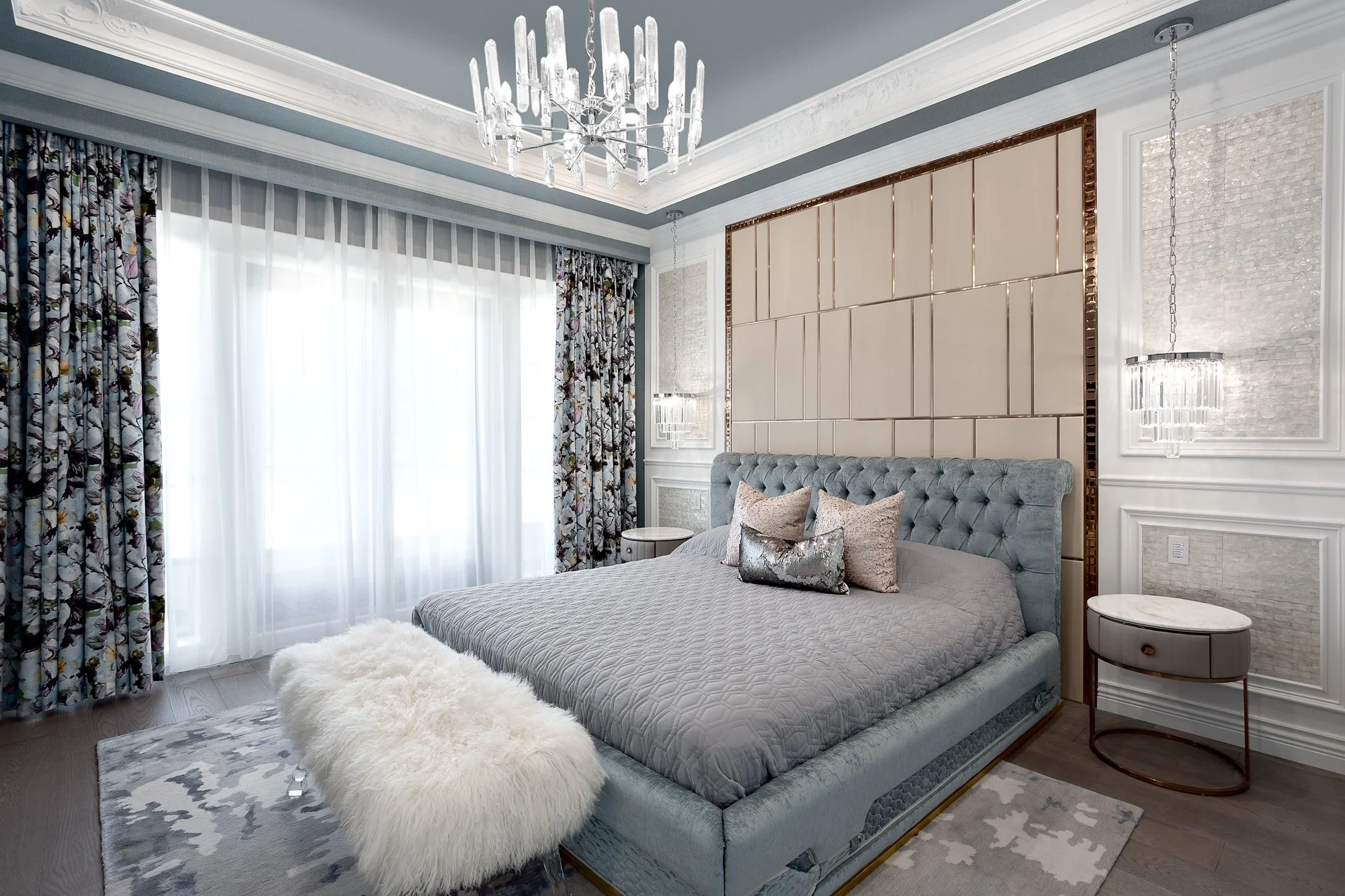 Luxury bedroom featuring a tufted blue bed frame, a custom tan paneled headboard wall, and a crystal chandelier.