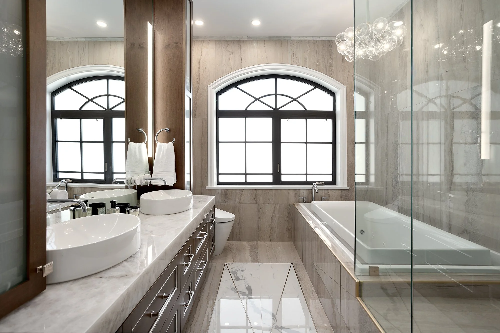 Sophisticated master bathroom with dual vanities, an arched window, and a built-in tub surrounded by white marble.