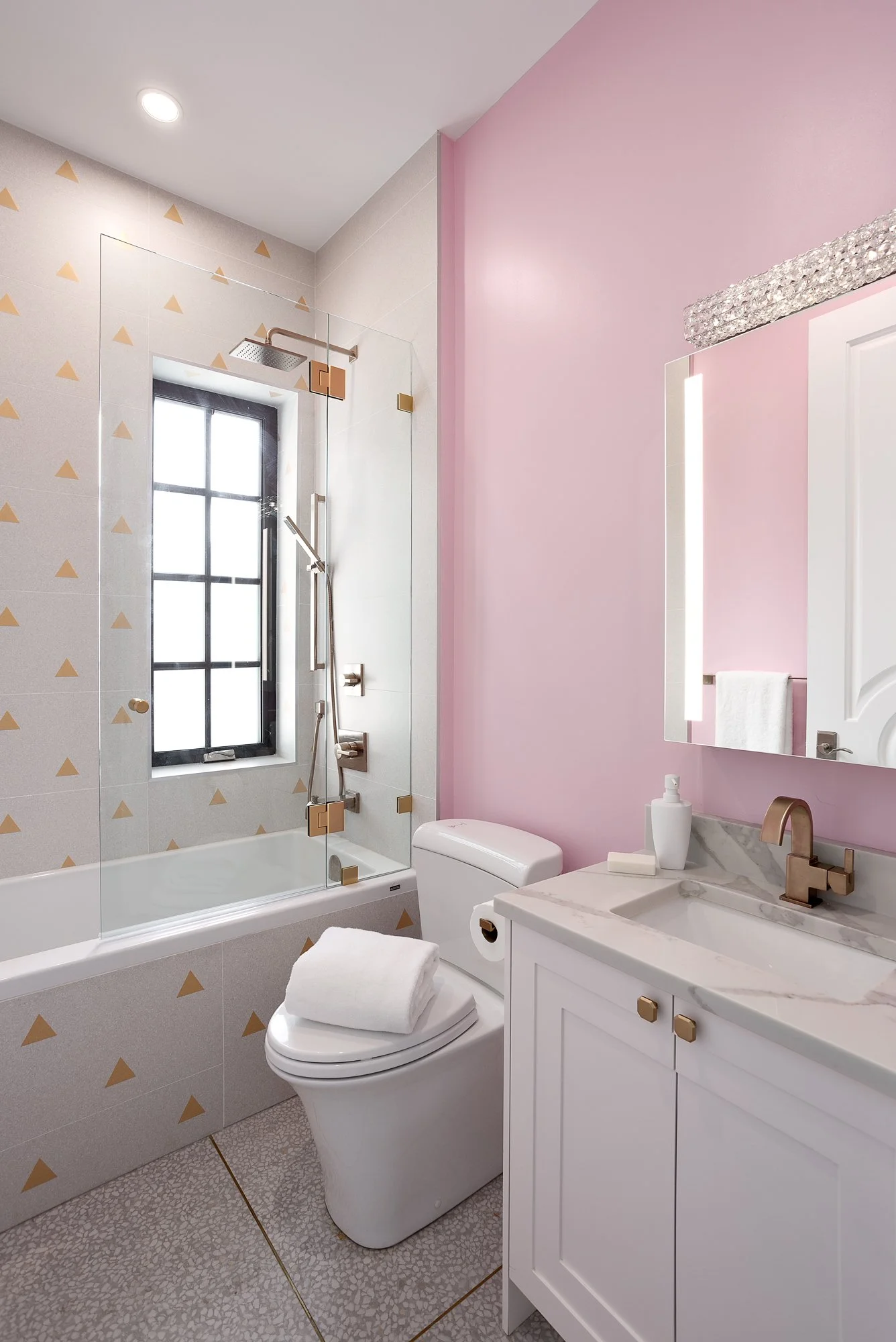 Playful modern bathroom with soft pink walls, a white vanity with gold hardware, and a geometric-patterned shower wall.