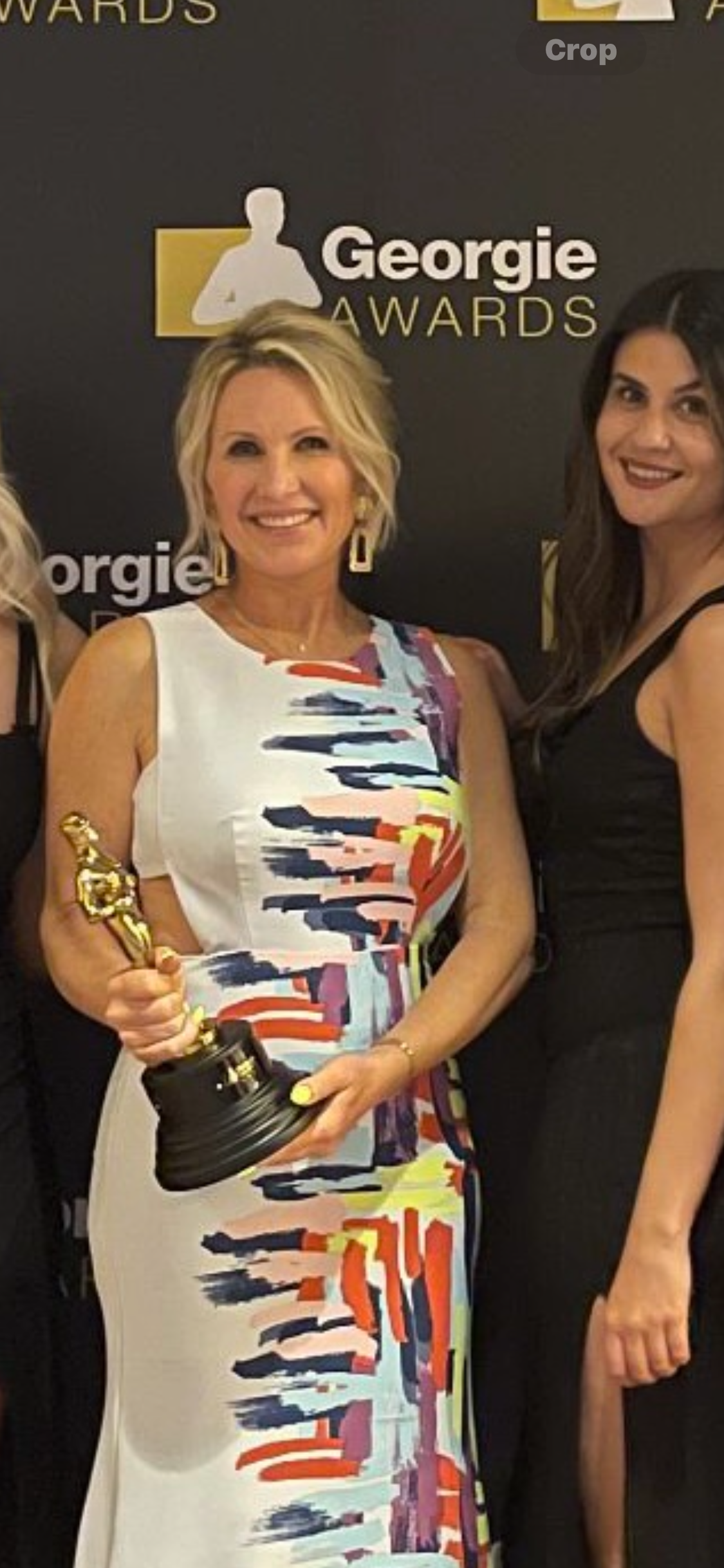 Woman holding an award at the Georgie Awards event, standing next to a woman in a black dress, with a black backdrop featuring the Georgie Awards logo.