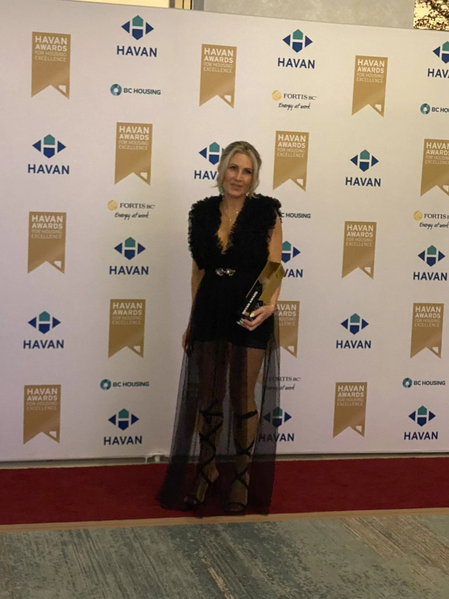 A woman in a black dress with sheer skirt and black high heels is standing on a red carpet in front of a step and repeat backdrop with logos for HAVAN and BC Housing, at an awards event.
