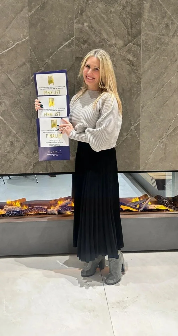A woman with blonde hair smiling and holding awards labeled "FINALIST" in front of a wall and a fireplace.