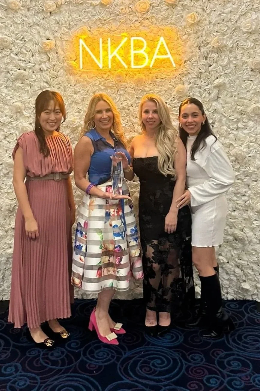Four women standing in front of a white floral wall with a neon sign that reads "NKBA." One woman is holding a clear award. The women are dressed in colorful and elegant outfits, smiling for the photo.