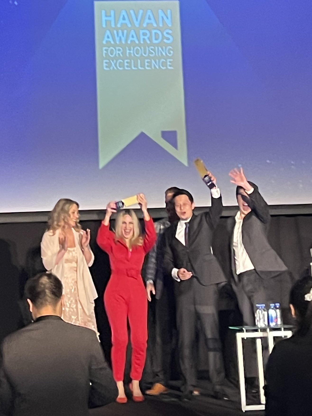 Group of four people celebrating on stage at the Havan Awards for Housing Excellence, with a large banner in the background and water bottles on a table in front.